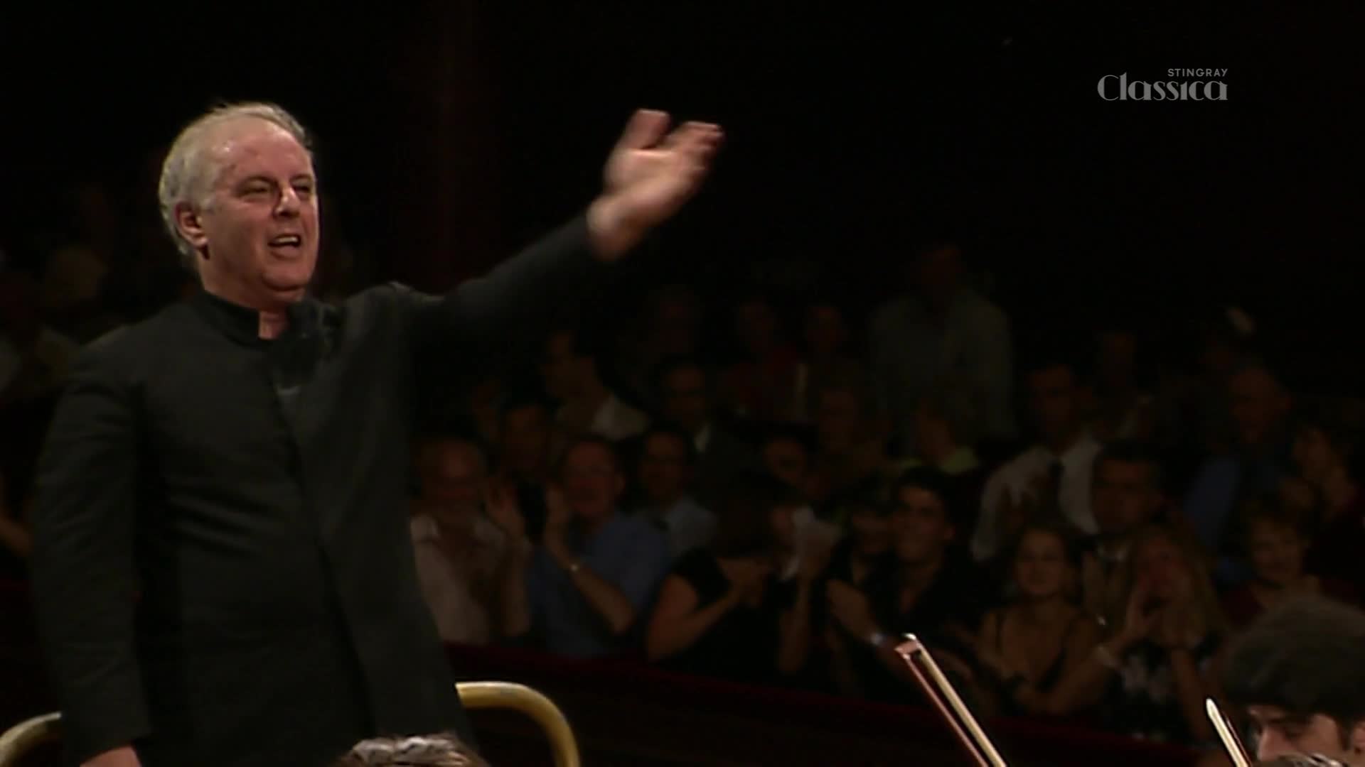 The conductor, in a black suit, raises his right arm, directing the orchestra. Behind him, the audience watches intently, the Stingray Classica logo visible in the corner.
