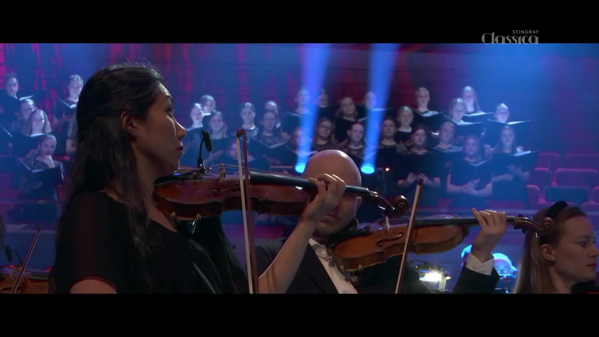 Violinists bow their instruments in unison, their faces focused on the music. Behind them, a choir stands in tiered rows, illuminated by spotlights from above.
