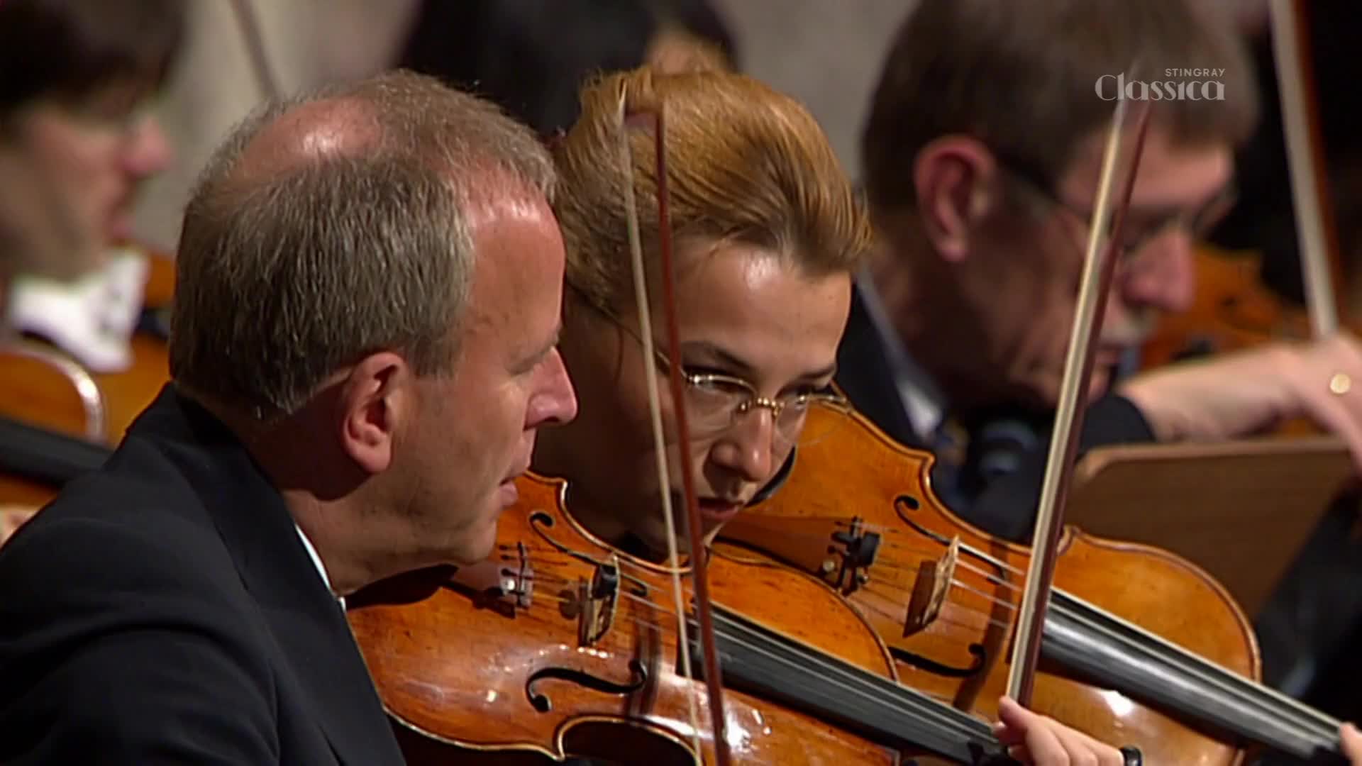 A violinist in glasses focuses intently on the music, her bow moving across the strings. Beside her, a man with a receding hairline leans in, also playing his instrument.
