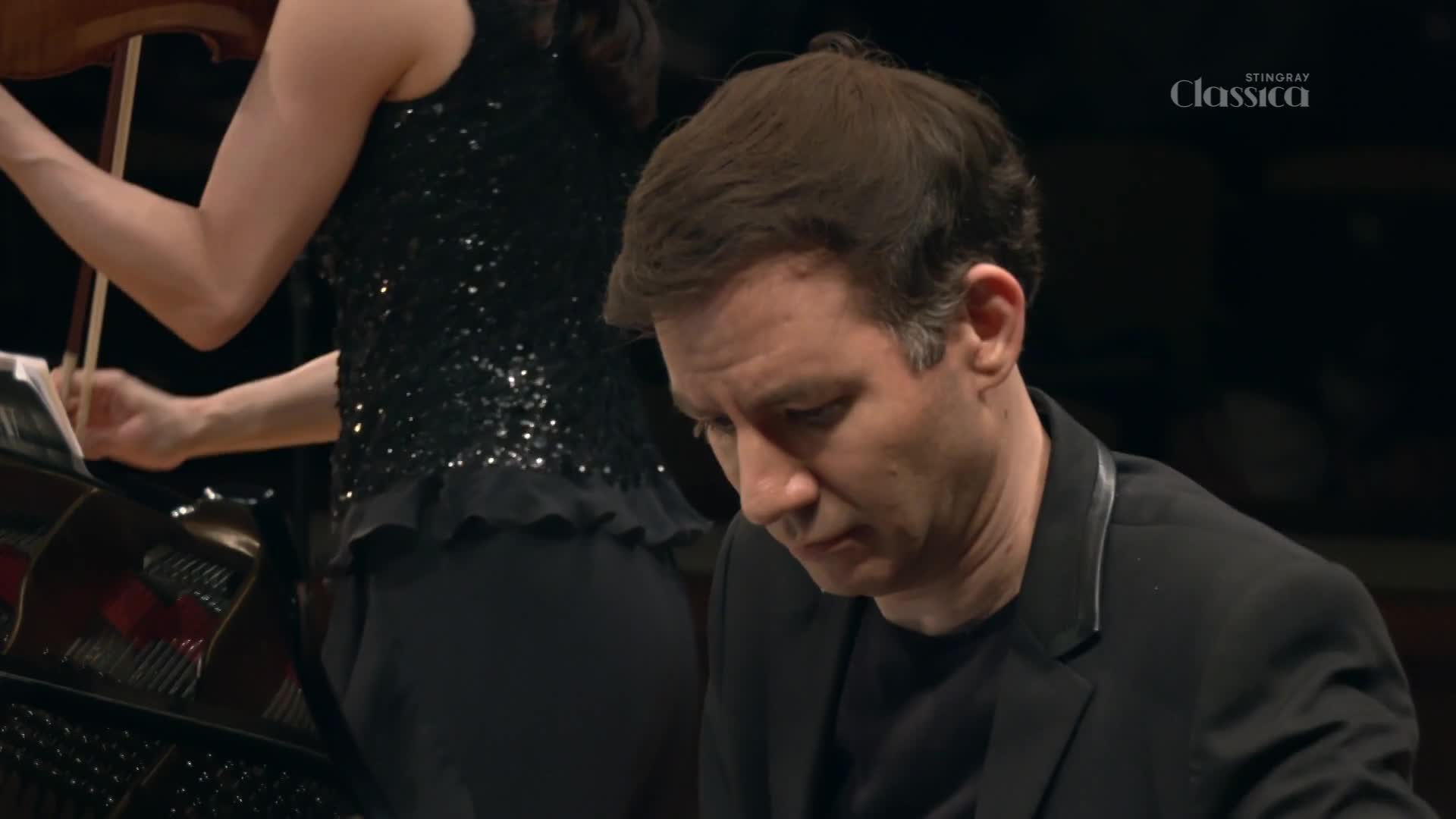 The pianist's hands are out of view, but his focused expression says everything. A violinist, in a shimmering top, stands at the ready beside him.
