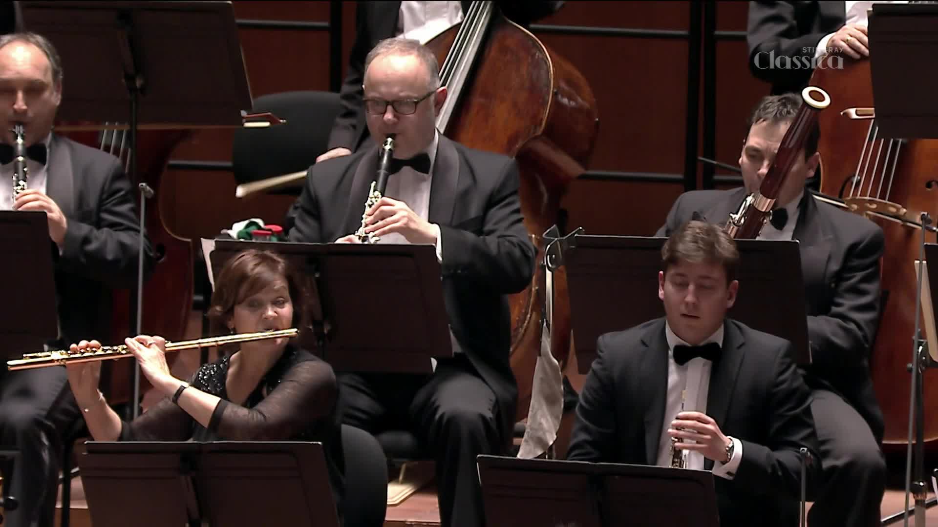 The woodwinds are in full focus, their instruments raised as they play. The musicians, dressed in formal attire, are intently focused on their sheet music during the performance.
