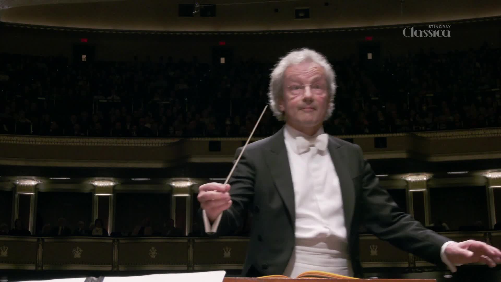 A conductor, dressed in a tuxedo, holds a baton aloft, his expression focused. The audience, dimly lit, sits in tiered rows behind him.
