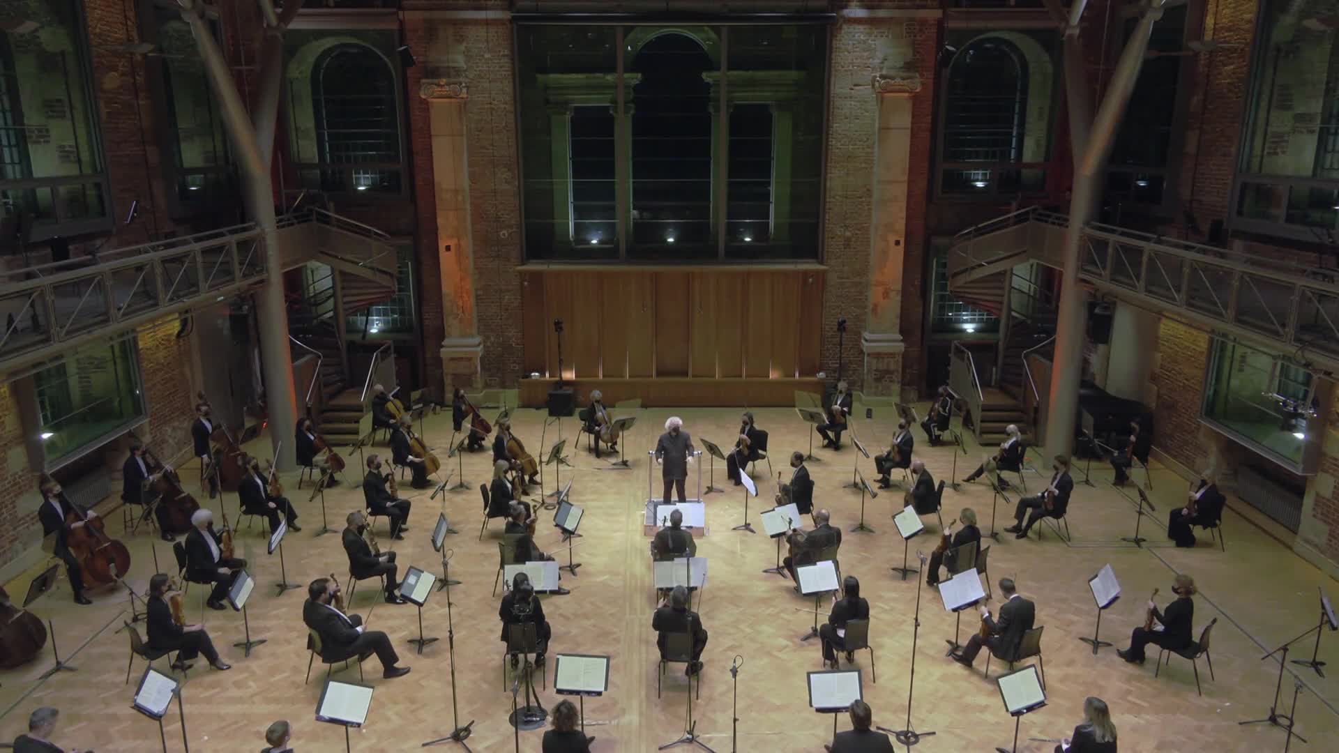 An orchestra, spread out across the floor, is poised, instruments at the ready. The conductor, with a shock of white hair, raises his baton, and the music is about to begin.
