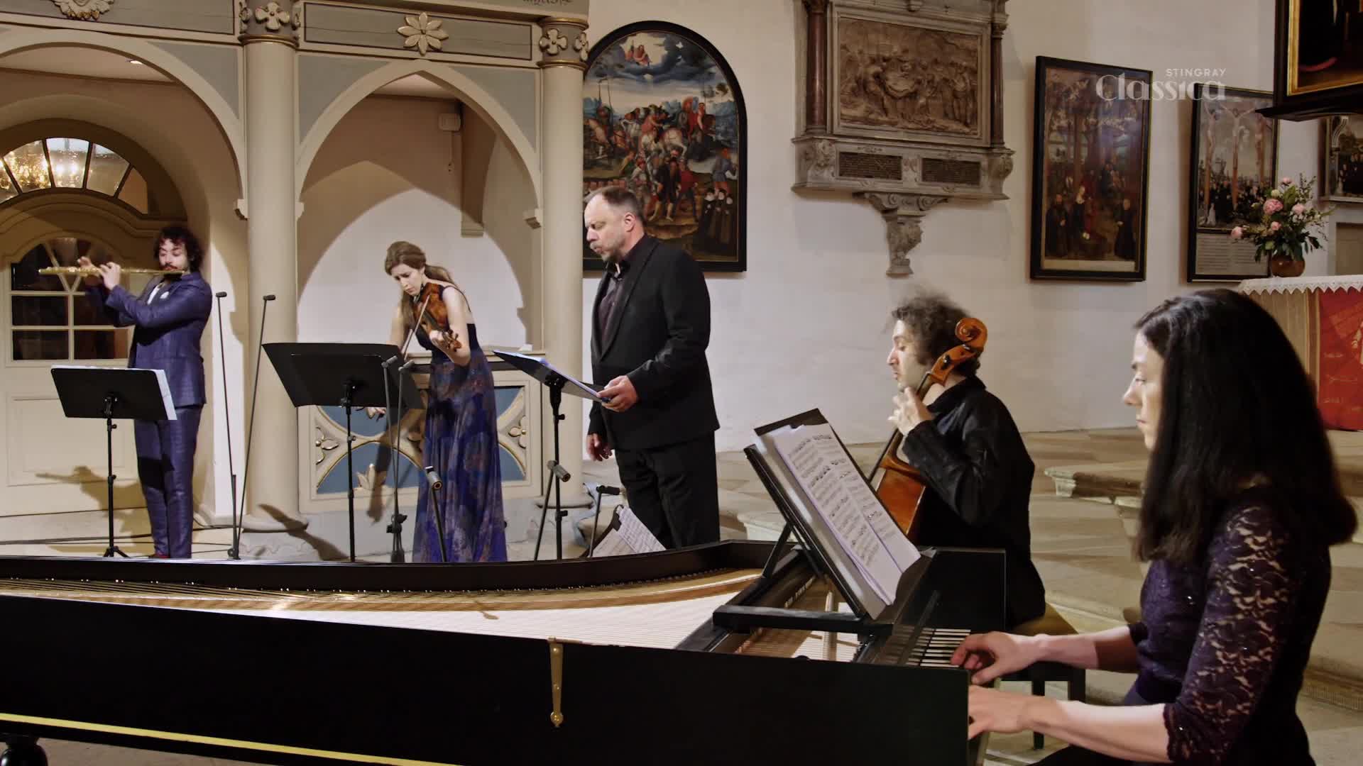 A small ensemble performs in a church, the flutist's breath visible as he plays. The woman at the piano's fingers dance across the keys, while the cellist bows his instrument.
