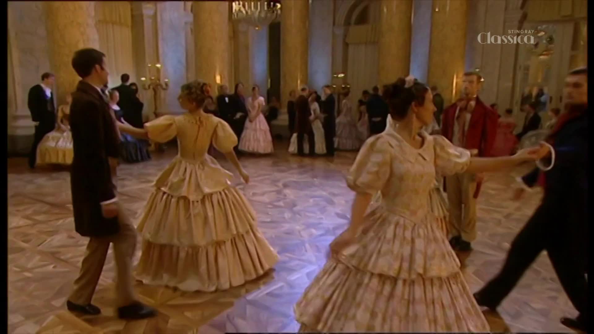 Couples in elaborate gowns and suits glide across the polished floor, their movements graceful and synchronized. The scene, broadcast on Stingray Classica from Canada, is a grand ballroom filled with dancers.
