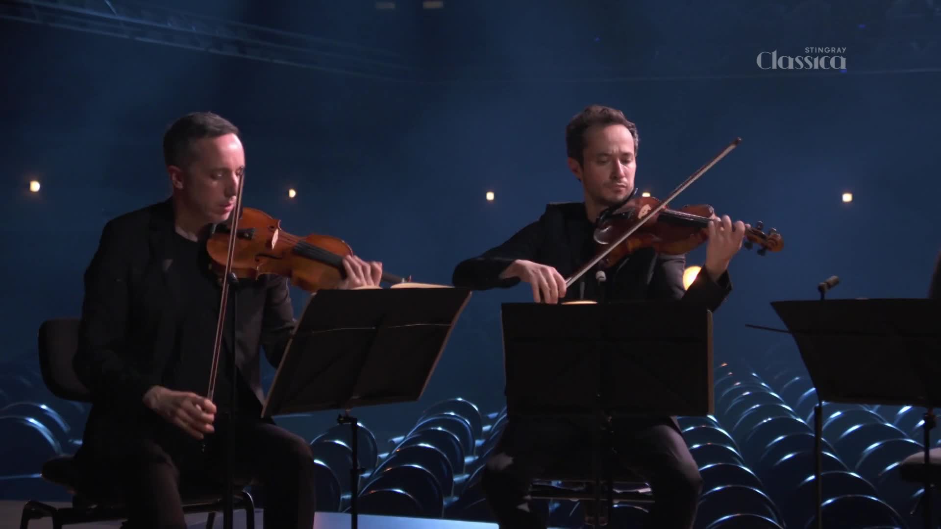 Two violinists, dressed in black, bow their instruments in unison, their movements precise and focused. The Stingray Classica logo is visible, and the stage is lit with a soft, blue glow.
