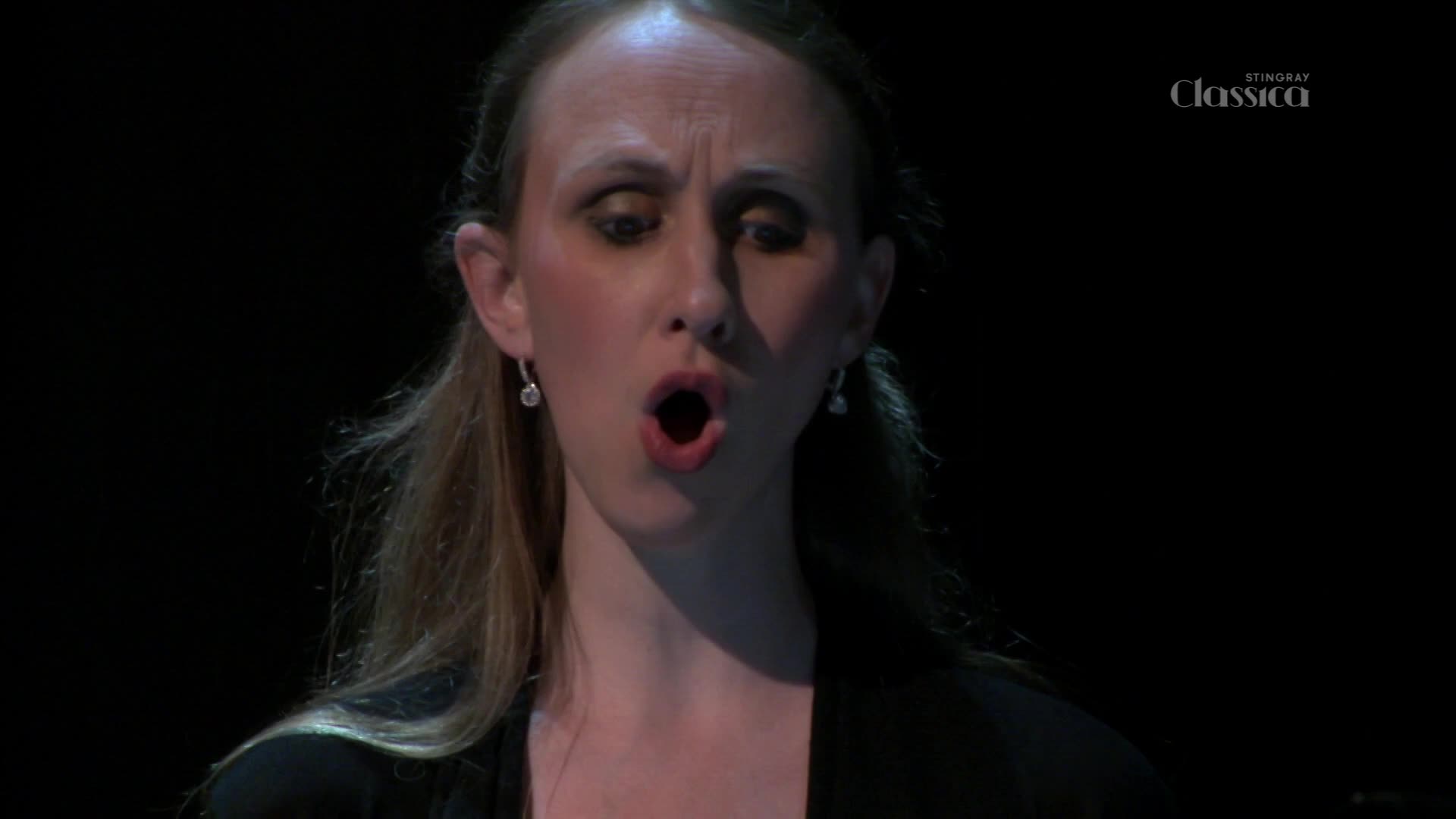 The woman's mouth is wide open, her face contorted as she sings on a Stingray Classica broadcast from Canada. Her eyes are fixed, and the dark background contrasts with her pale skin.
