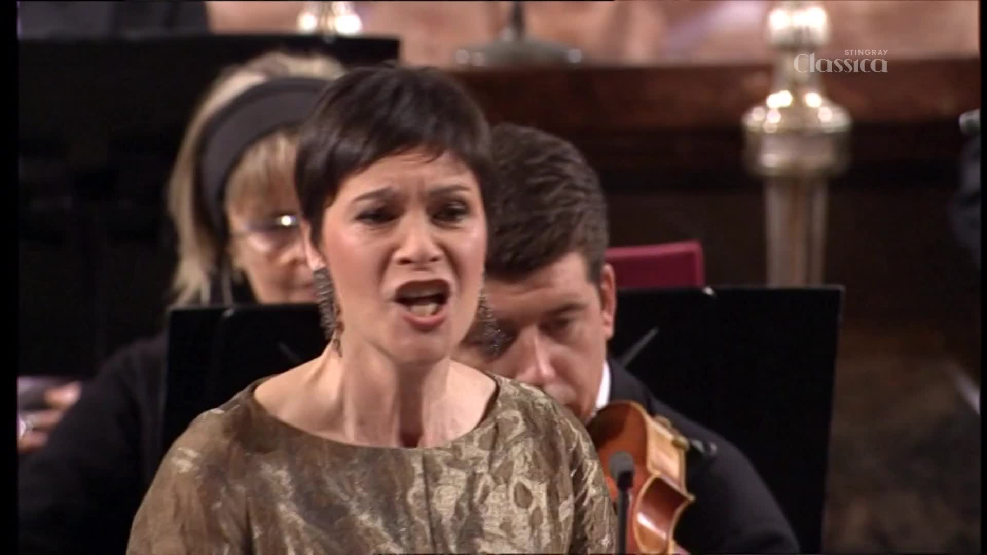 A woman in a shimmering gold dress is singing with her mouth wide open, her face showing intense emotion. Behind her, a violinist is focused on his instrument, part of the orchestra performing for Stingray Classica, a Canadian broadcast.
