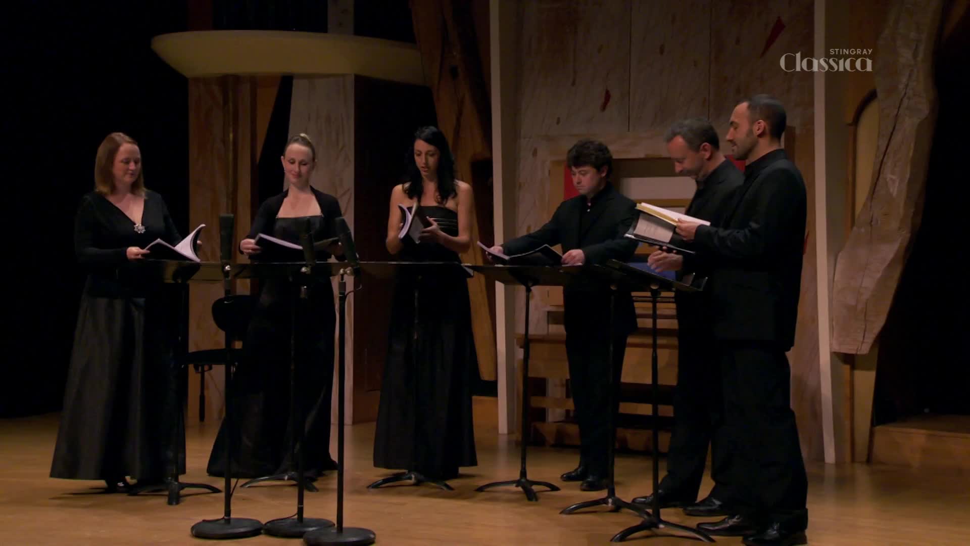 Six people in formal attire stand on a stage, each holding sheet music. They face forward, ready to perform for Stingray Classica, a Canadian broadcast.
