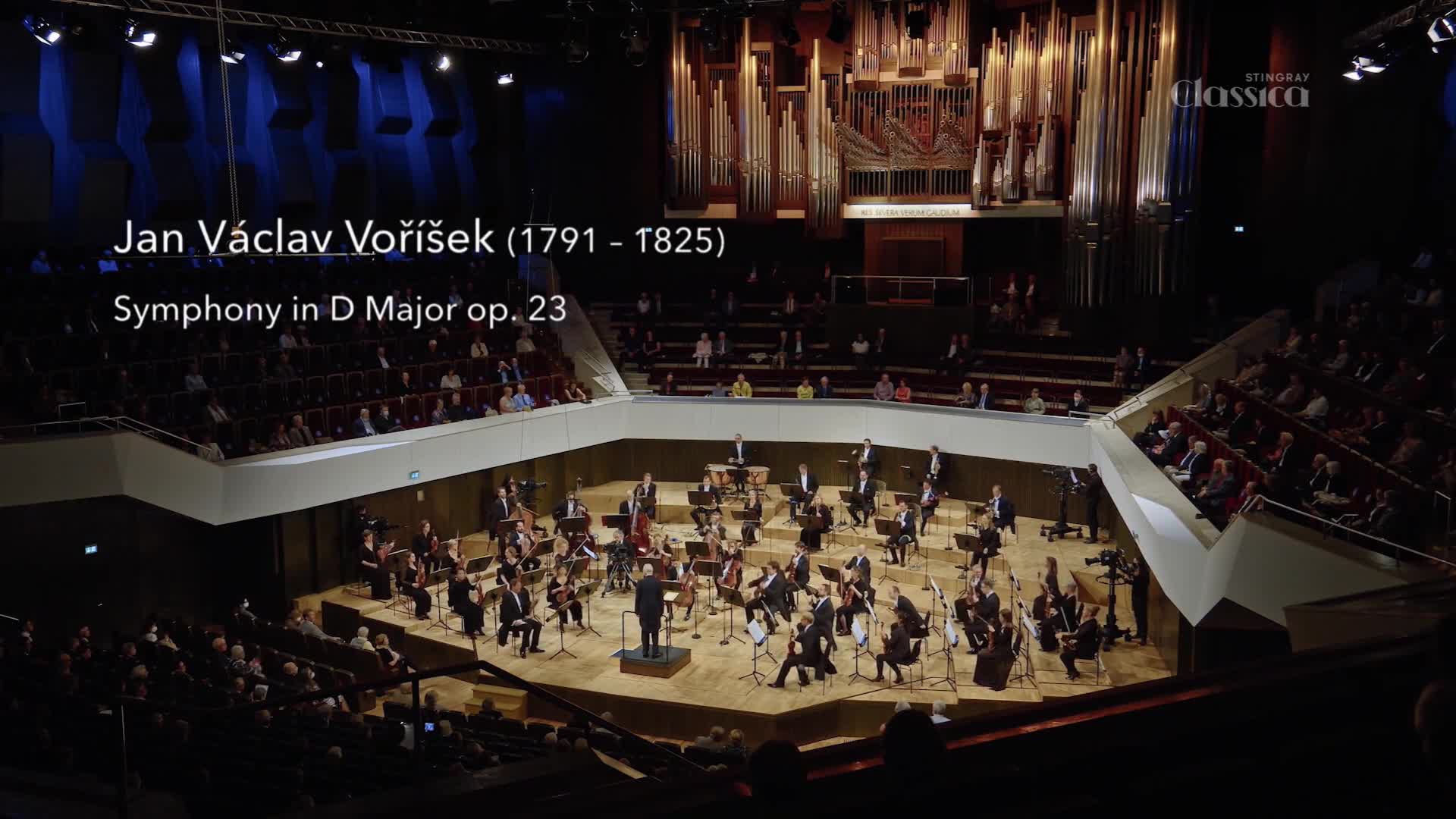 The conductor's baton slices through the air, guiding the orchestra in a performance broadcast by Stingray Classica. The musicians, in formal attire, are focused on their instruments within the concert hall.
