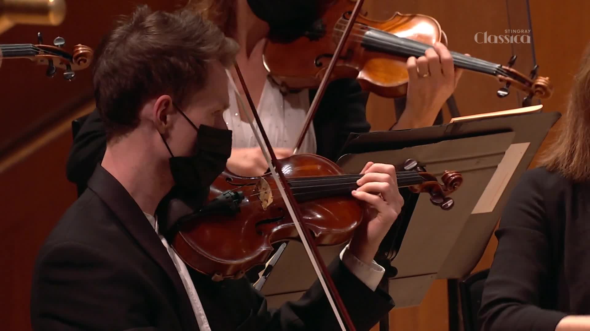 A violinist, masked and in a suit, draws his bow across the strings. The camera angle is close, capturing the fine movements of his fingers and the instrument.
