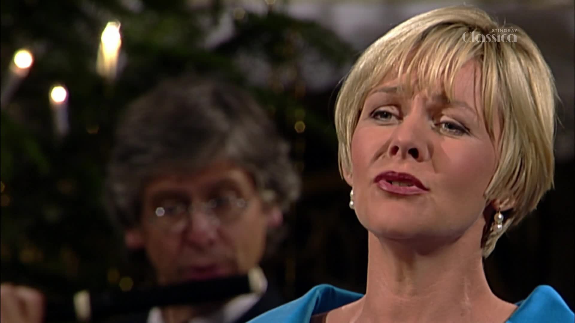 A woman with short blonde hair sings, her lips moving as she performs. Behind her, a man with glasses plays a flute, his breath visible as he blows.
