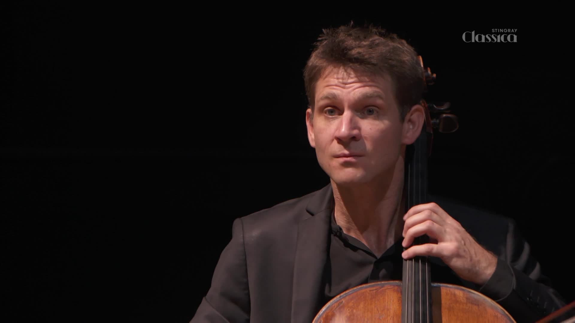 The cellist, dressed in a dark suit, holds his instrument, his expression focused. The Stingray Classica logo is visible, suggesting a performance is being broadcast from Canada.
