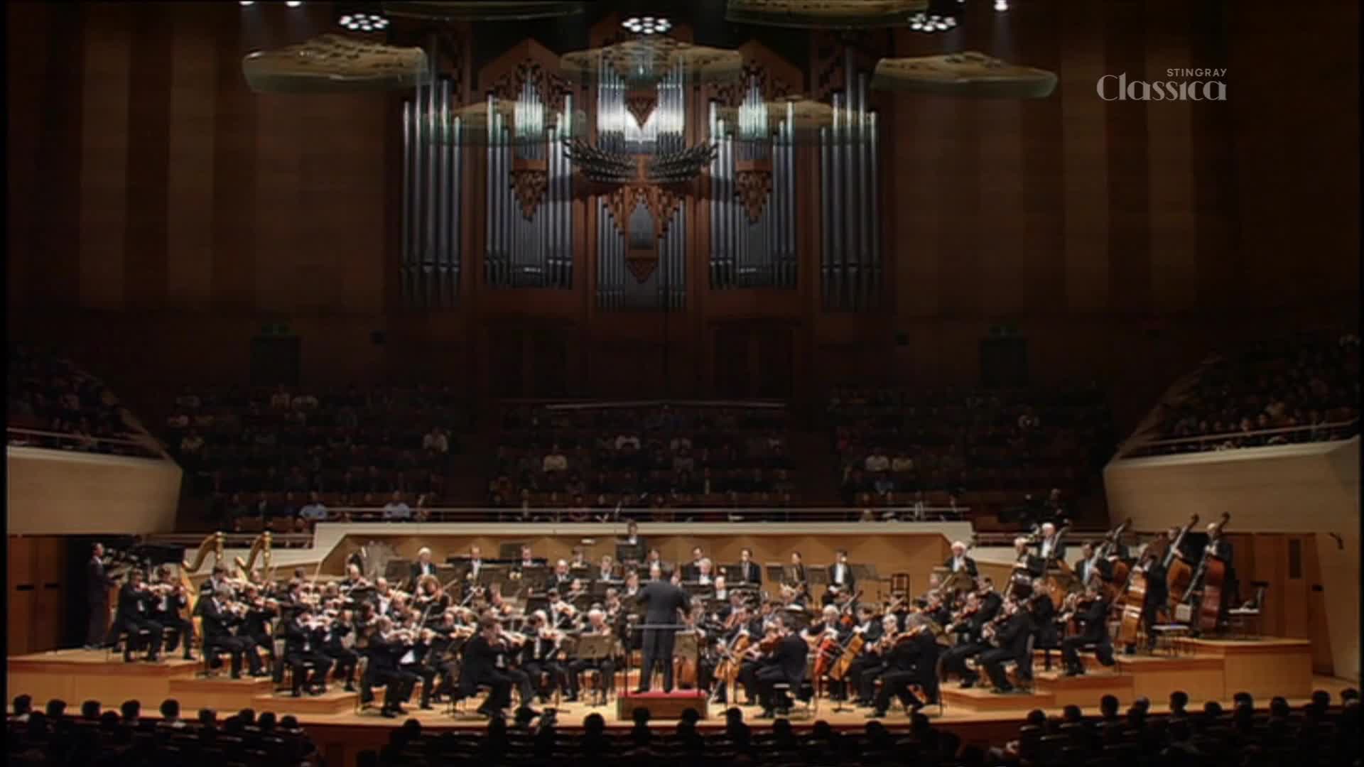The conductor's baton moves in broad strokes, and the orchestra responds, filling the hall with sound. The broadcast logo for Stingray Classica is visible, likely from Canada.
