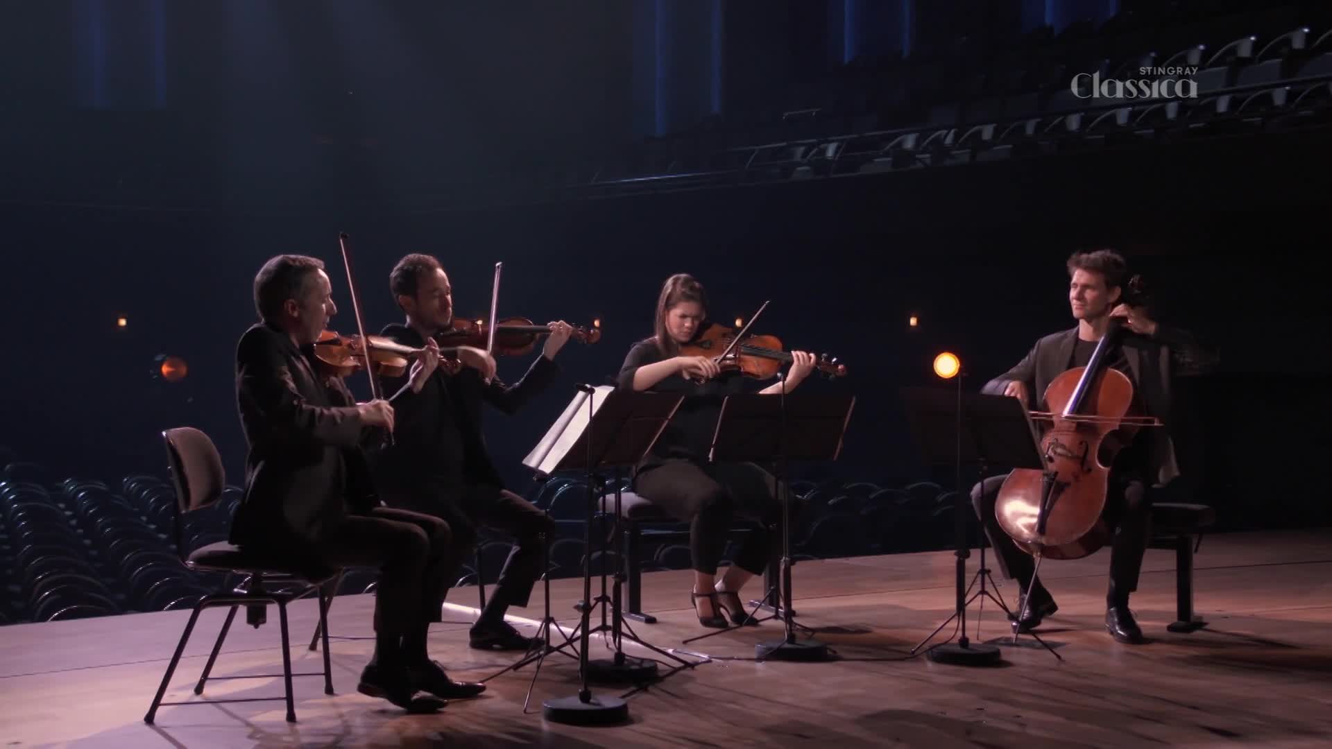 Four musicians are seated on a stage, bows moving across strings in a performance for Stingray Classica. The cellist's instrument is large and brown, contrasting with the black outfits of the violinists.

