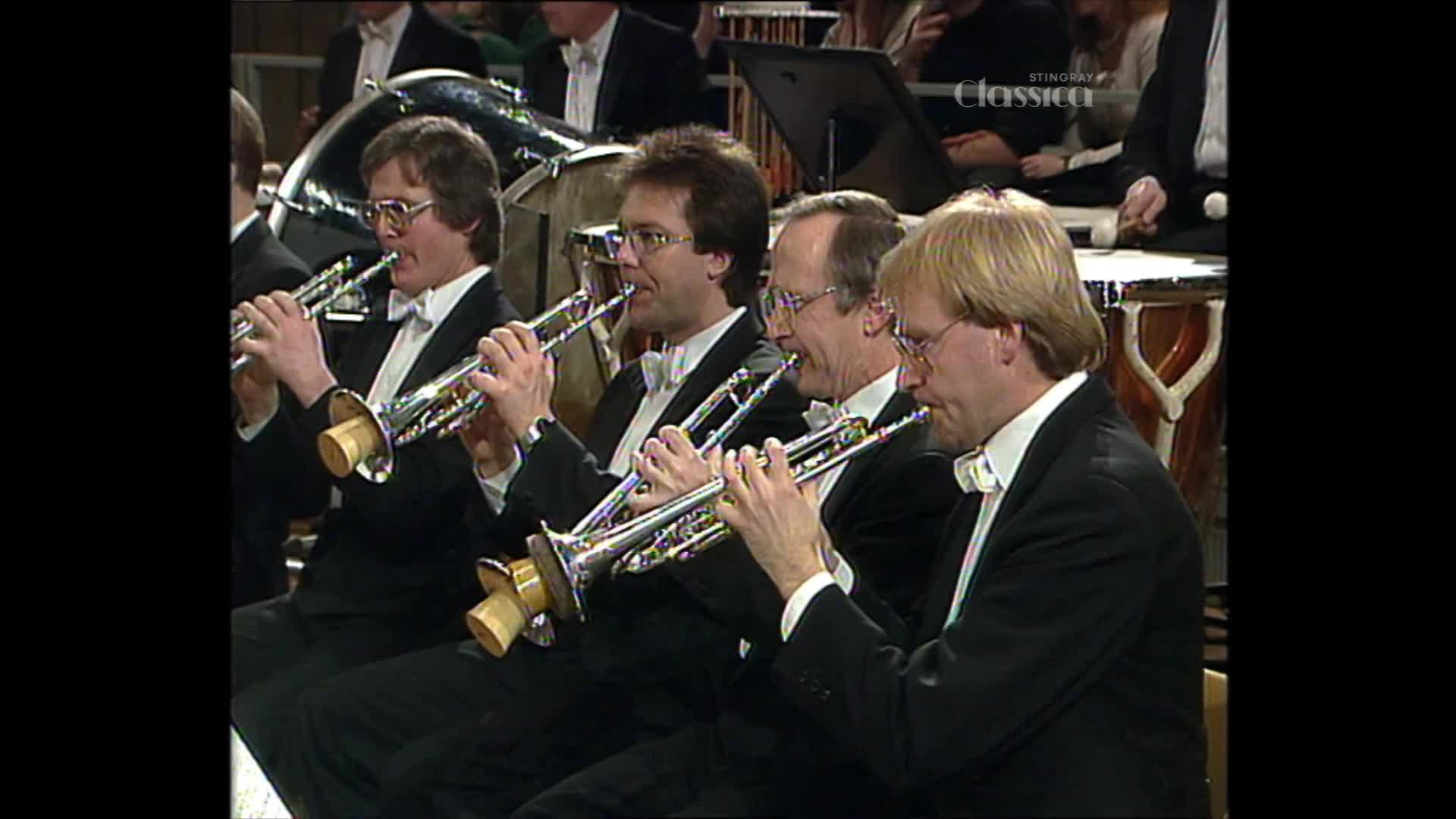 Four men in tuxedos are playing trumpets in unison. The polished brass instruments gleam under the stage lights as they create music.
