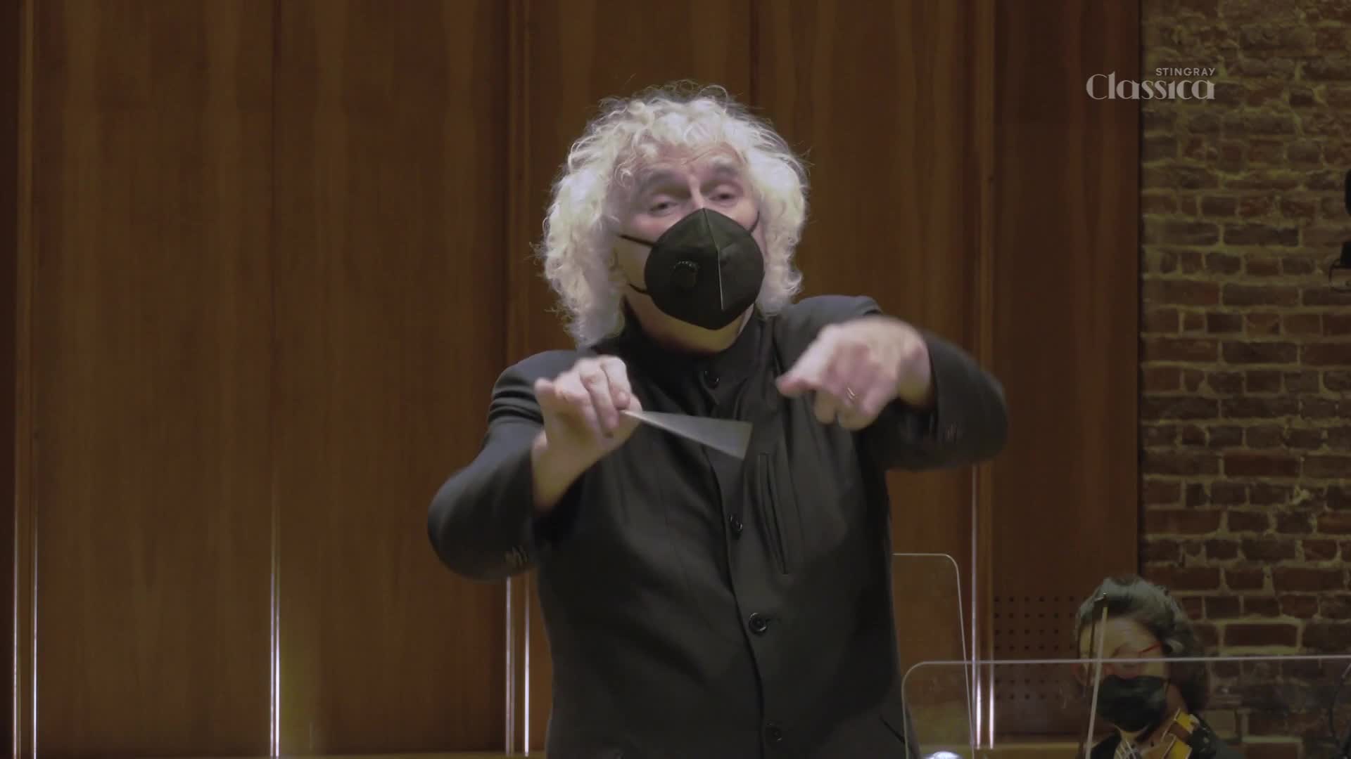 The conductor, his wild white hair framing a masked face, gestures with a baton, leading the Stingray Classica orchestra. He's in the middle of a passionate sweep, his left hand pointing forward as his right guides the baton in a sharp, decisive arc.