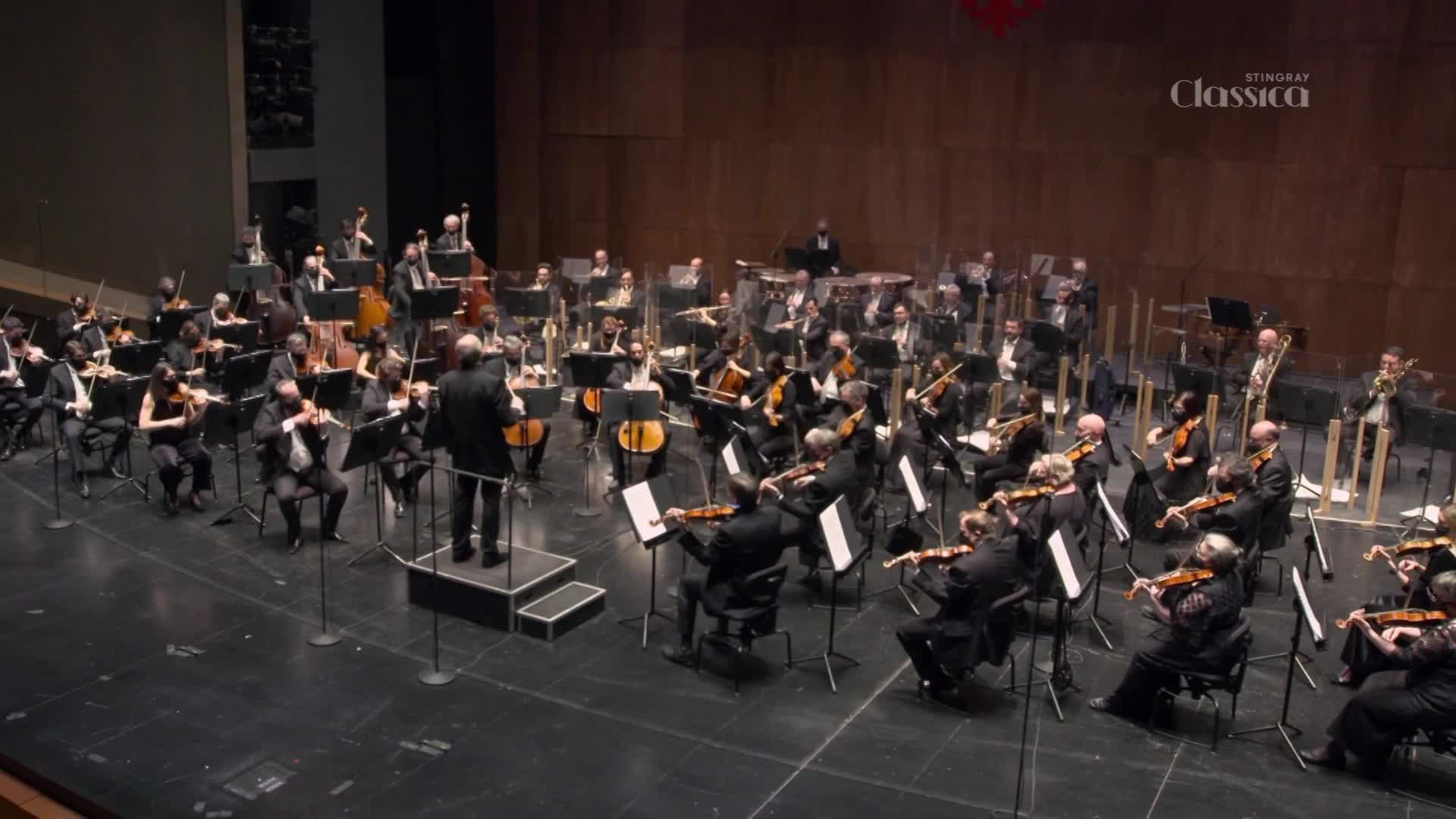 The conductor stands on a raised platform, leading a full orchestra as they play their instruments. The Stingray Classica logo is visible in the upper right corner of the stage. The conductor stands on a raised platform, leading a full orchestra as they play their instruments. The Stingray Classica logo is visible in the upper right corner of the stage.