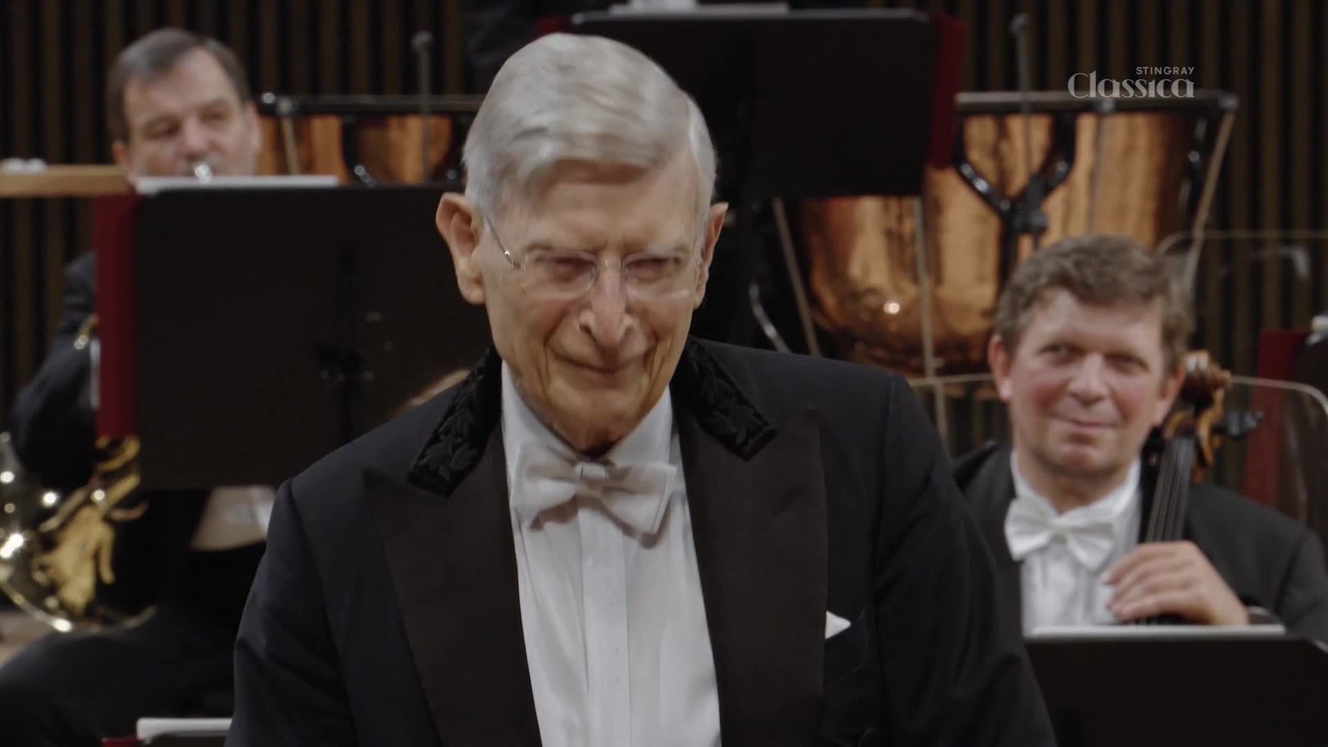 The conductor, dressed in a tuxedo, smiles warmly as he stands before the orchestra. A cellist in the background, also in formal wear, returns his gaze with a friendly expression. The conductor, dressed in a tuxedo, smiles warmly as he stands before the orchestra. A cellist in the background, also in formal wear, returns his gaze with a friendly expression.