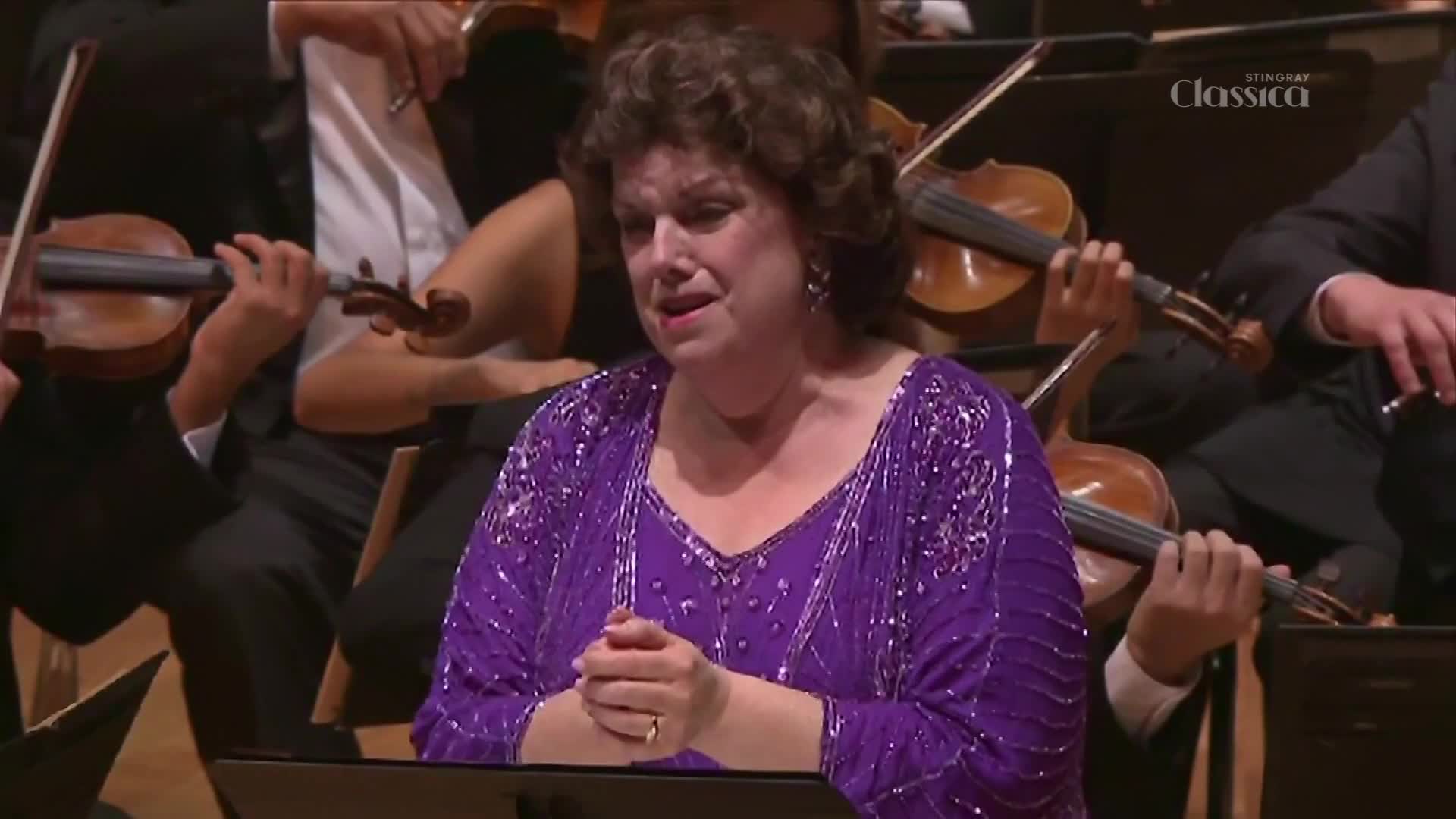 A woman in a sparkling purple gown sings with her hands clasped before her. Violinists in the orchestra behind her draw their bows across the strings. A woman in a sparkling purple gown sings with her hands clasped before her. Violinists in the orchestra behind her draw their bows across the strings.