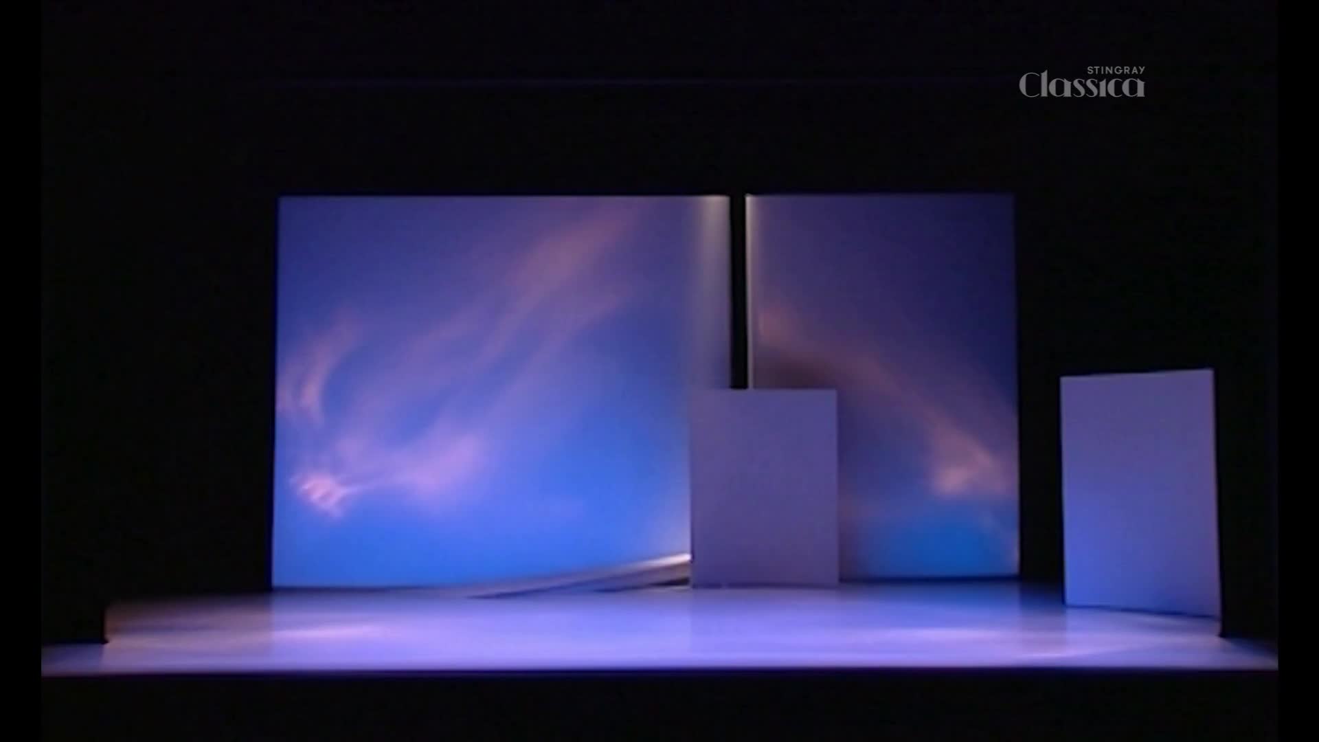 A stage is set with a large blue backdrop featuring swirling pinkish clouds. Three white rectangular blocks stand on the polished floor, catching the light. A stage is set with a large blue backdrop featuring swirling pinkish clouds. Three white rectangular blocks stand on the polished floor, catching the light.