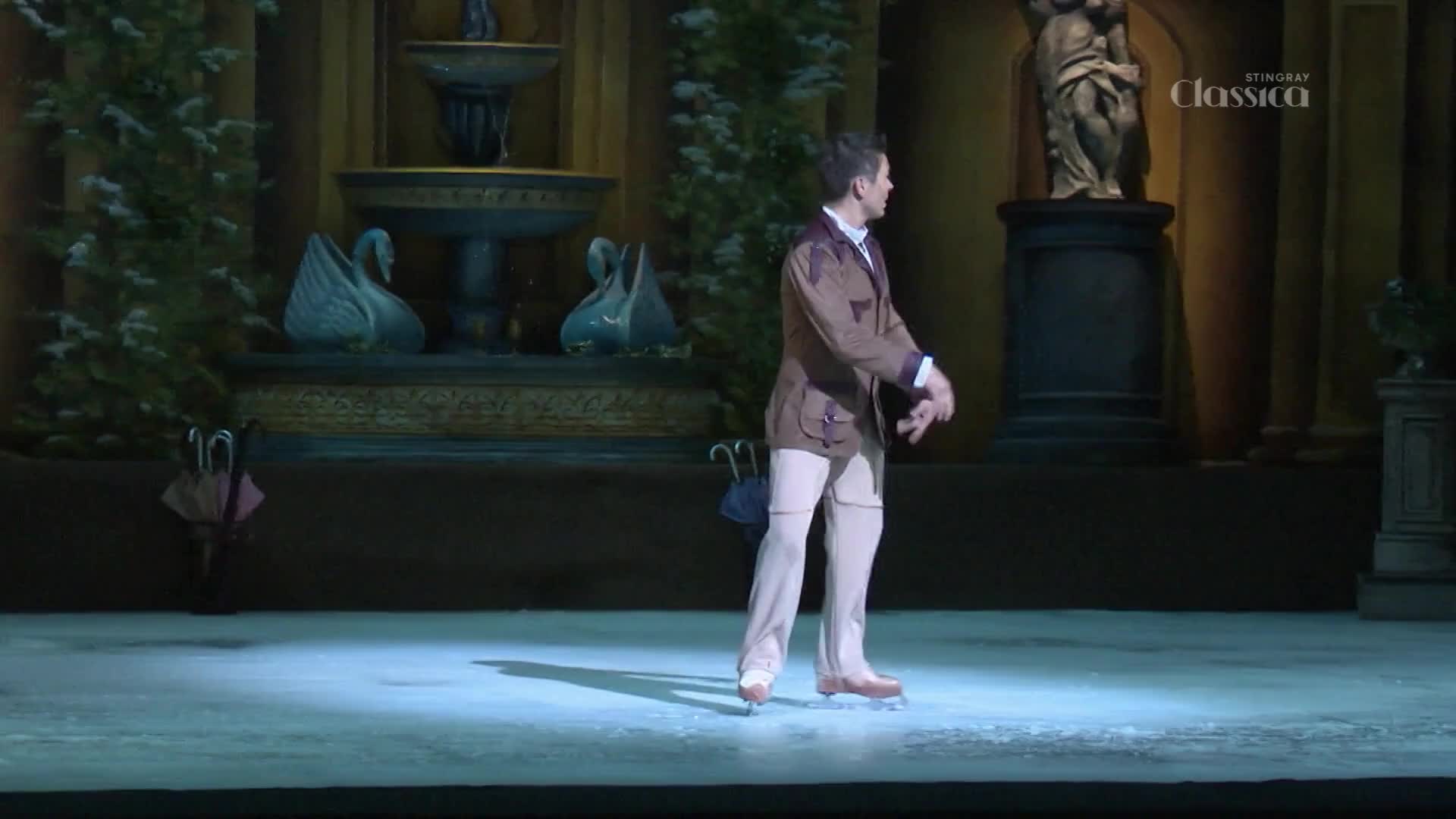 A lone figure in a brown jacket and light trousers glides across the ice, his arms outstretched. The stage behind him is set with a fountain and two swan sculptures, reminiscent of a grand Canadian garden. A lone figure in a brown jacket and light trousers glides across the ice, his arms outstretched. The stage behind him is set with a fountain and two swan sculptures, reminiscent of a grand Canadian garden.