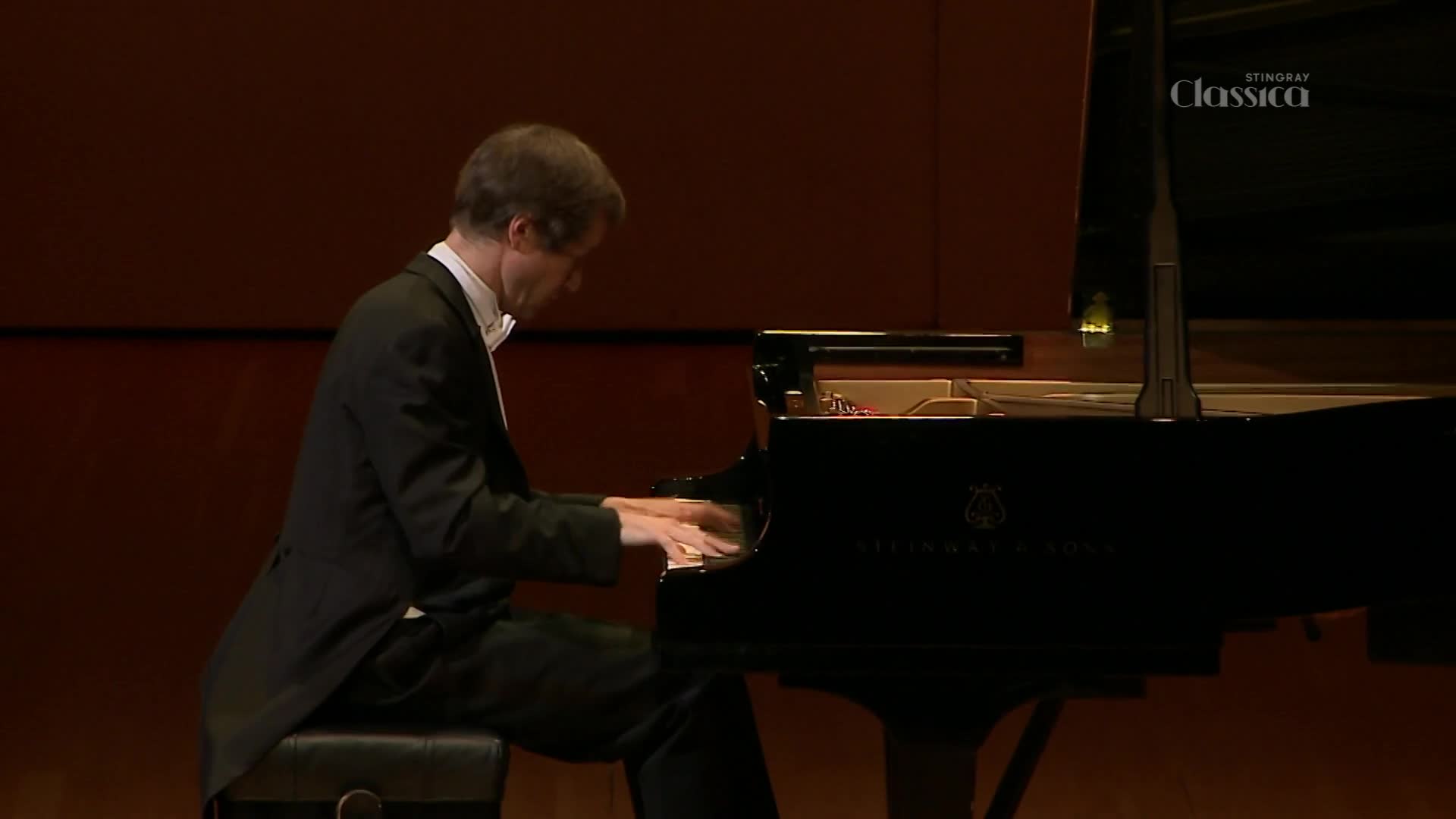 A man in a tuxedo plays a Steinway grand piano on a stage. The Stingray Classica logo is visible in the upper right corner. A man in a tuxedo plays a Steinway grand piano on a stage. The Stingray Classica logo is visible in the upper right corner.