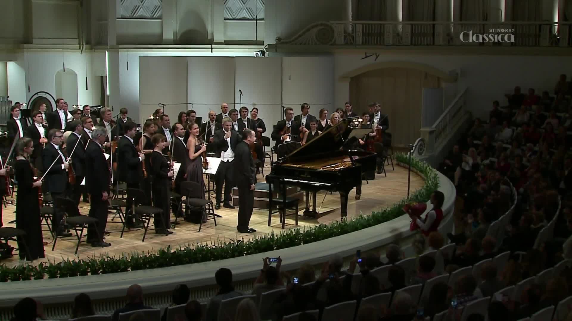 A pianist stands beside a grand piano as an orchestra prepares to play. A conductor, facing the musicians, gestures with his hands.