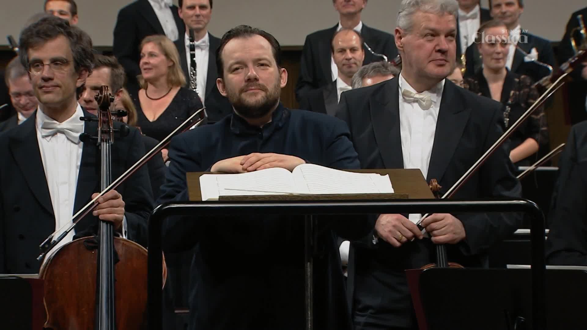 A cellist in a tuxedo stands ready, his bow poised over the strings. Beside him, a conductor rests his hands on an open score, a sea of musicians in formal wear behind them.