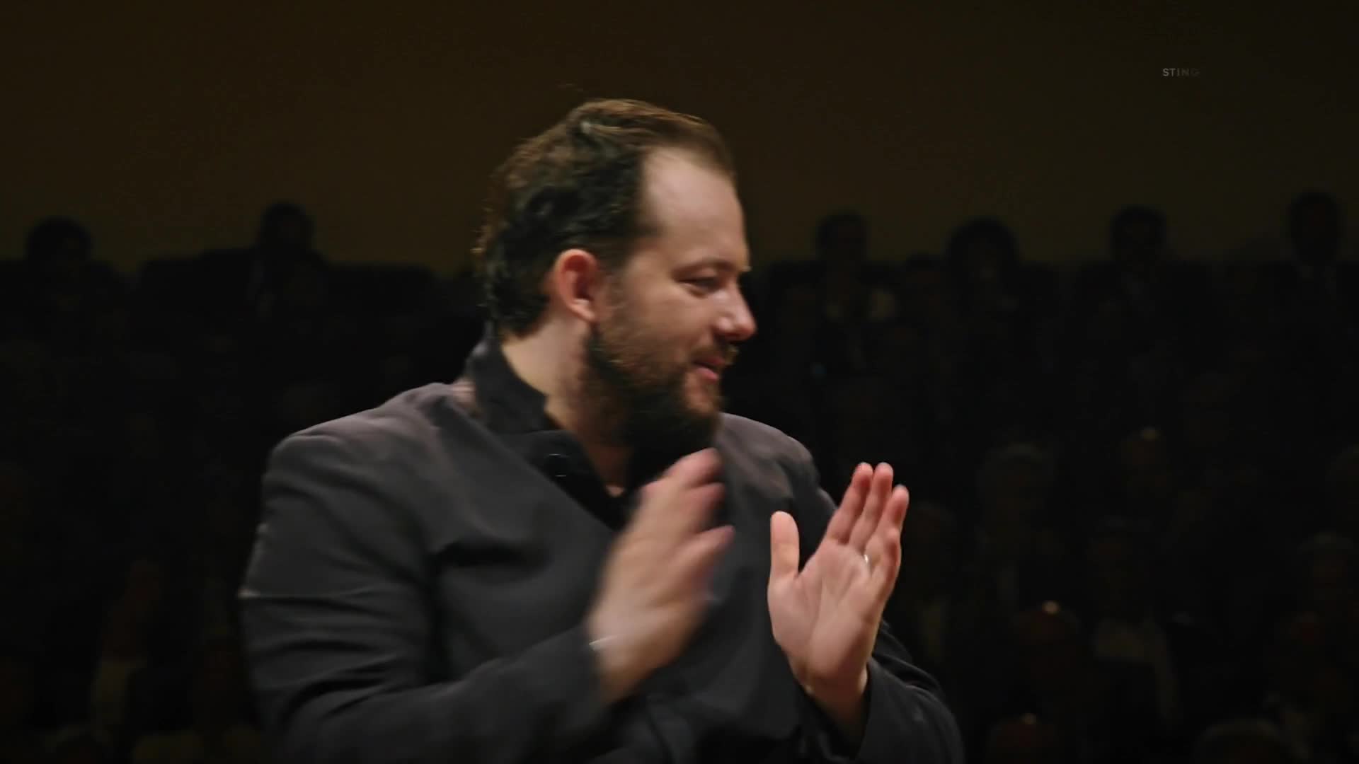 The conductor claps his hands together, a smile on his face. He stands on a stage, facing an audience in the dim light.