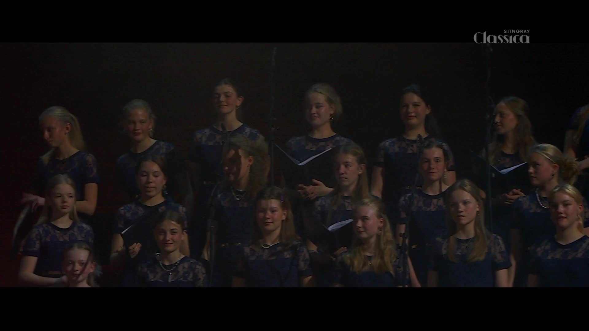 A group of young women in matching navy dresses stand in rows, some holding sheet music. The Stingray Classica logo appears in the upper right corner.