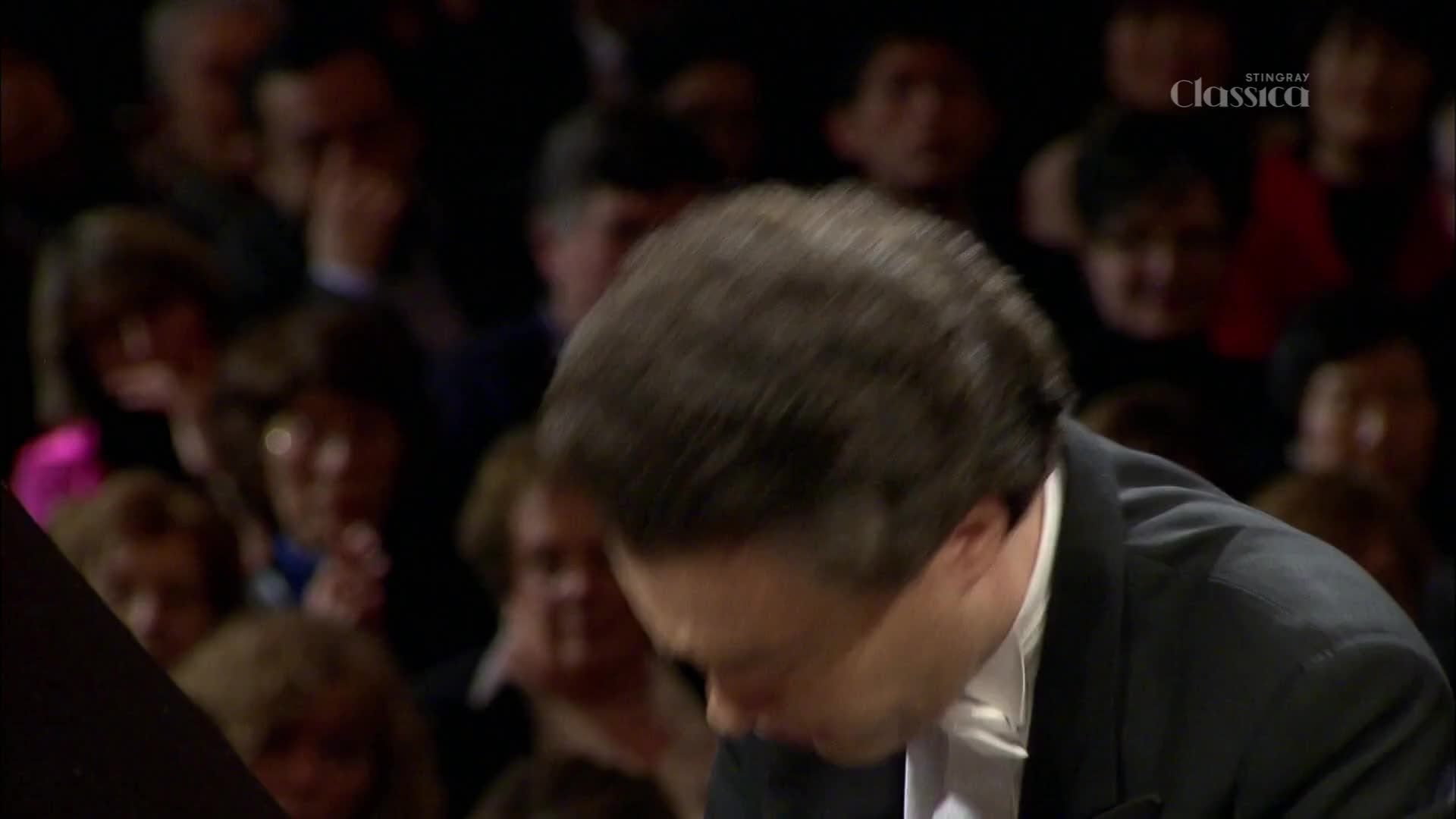 The pianist's head bows low, his dark hair a blur as his hands move across the keys. The audience in the concert hall, seen through Stingray Classica, watches intently.