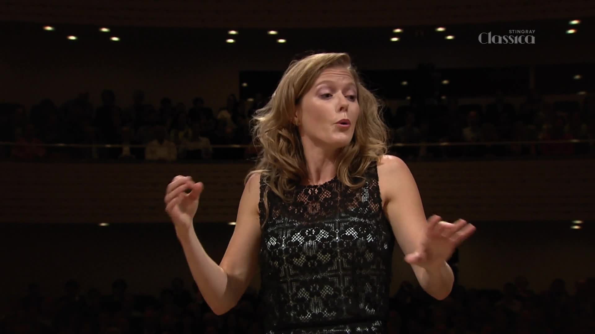 The conductor's hands move with purpose, guiding the orchestra through a piece broadcast on Stingray Classica. A hushed audience watches from the balcony in this Canadian hall.