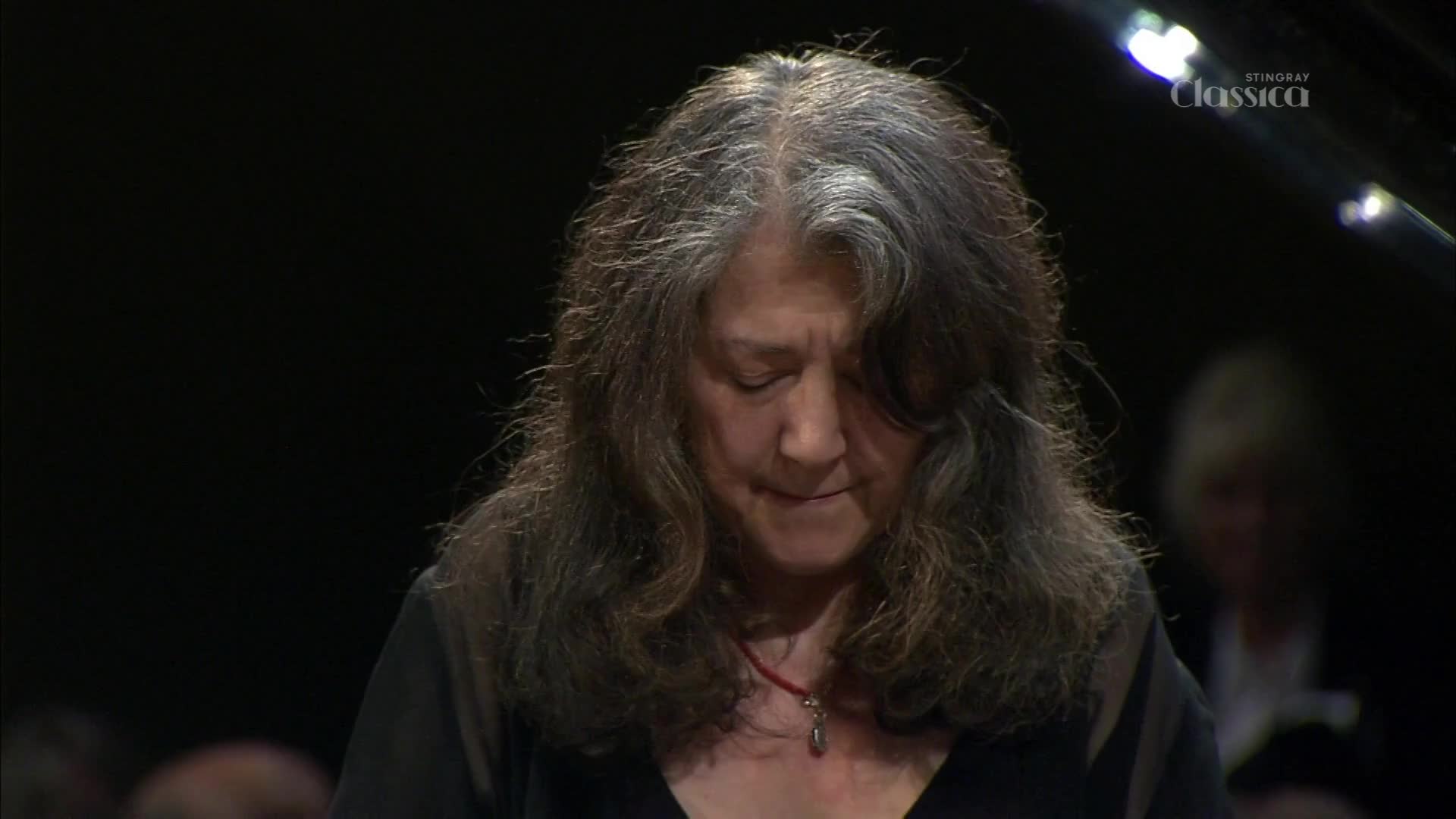 The pianist's head is bowed, her long, grey hair falling around her face.  A red pendant rests against her dark dress, catching the stage lights.