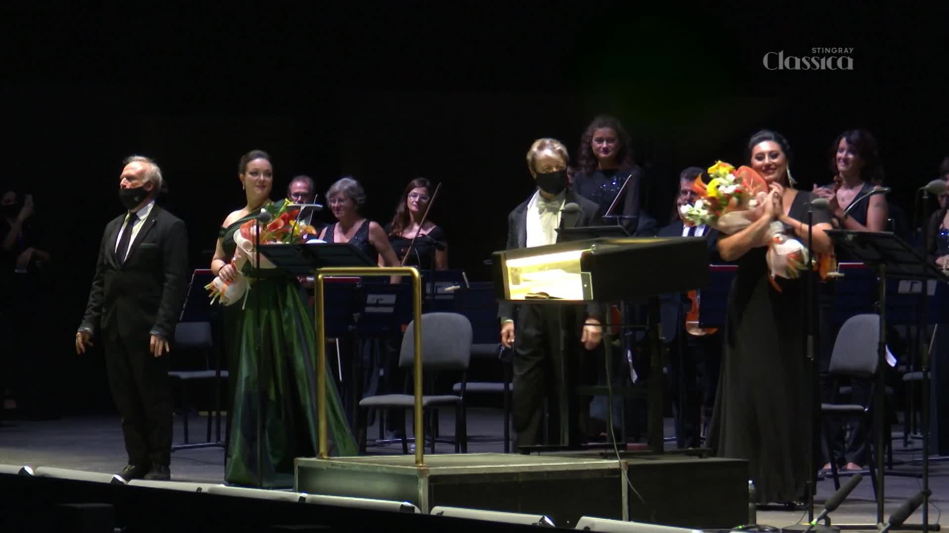 A man in a dark suit and a woman in a green gown stand center stage, holding bouquets. Behind them, an orchestra is gathered, some musicians holding instruments. The Stingray Classica logo appears in the upper right corner.