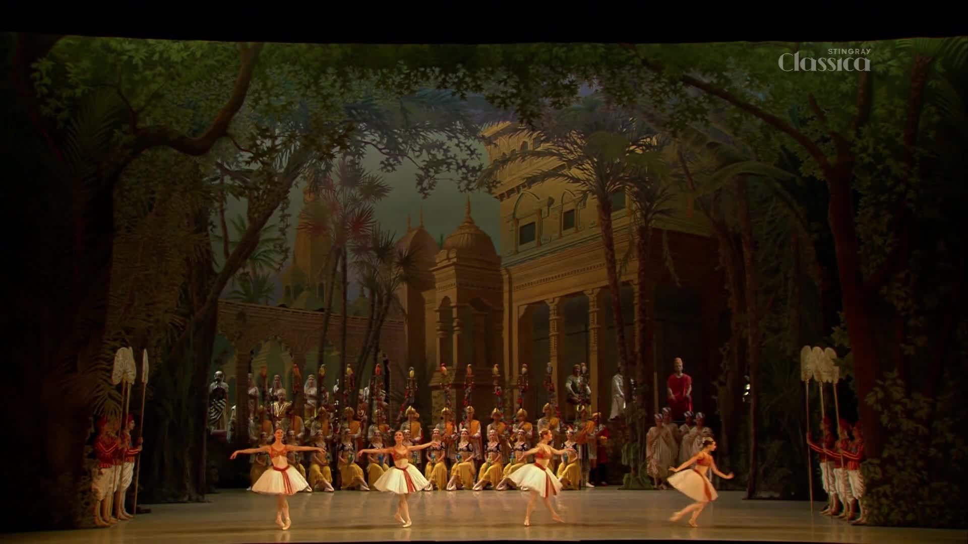 Dancers in white tutus and orange bodices glide across the stage. A backdrop depicts a lush, exotic landscape with ornate buildings, hinting at a performance from Canada's Stingray Classica.