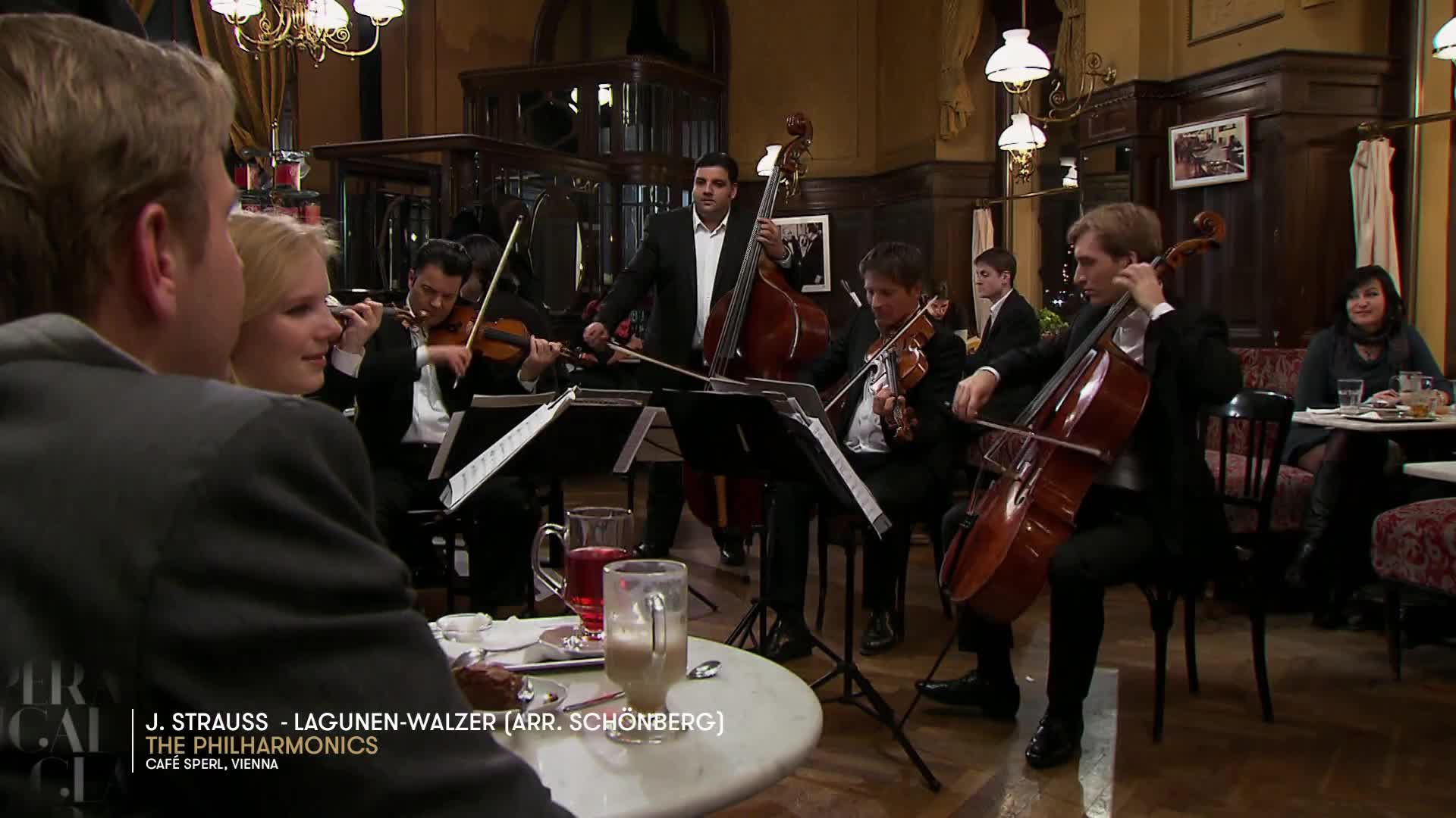 A cellist draws his bow across the strings, his instrument held close. Beside him, a violinist plays, her eyes focused on the music.