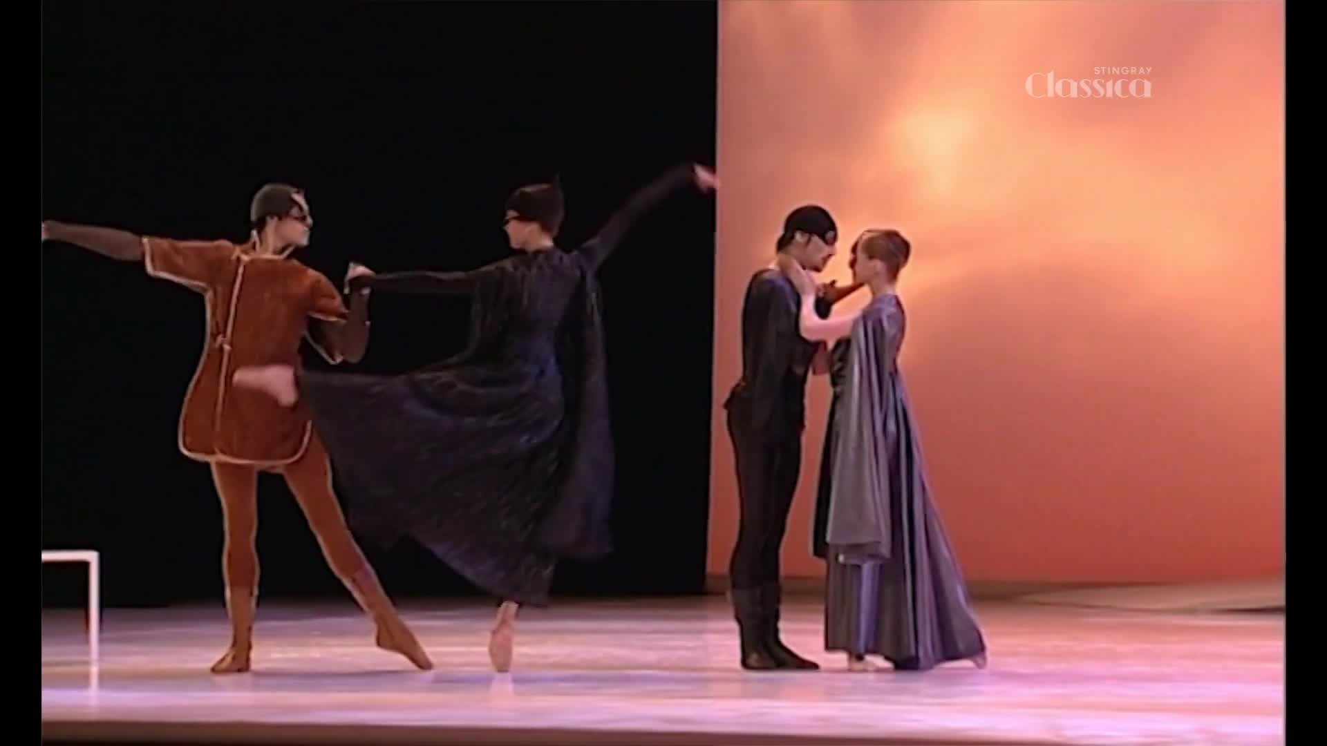 A dancer in a brown tunic and tights kicks out a leg while another in a dark cape pirouettes. Nearby, a couple in matching dark outfits embrace, their faces close.
