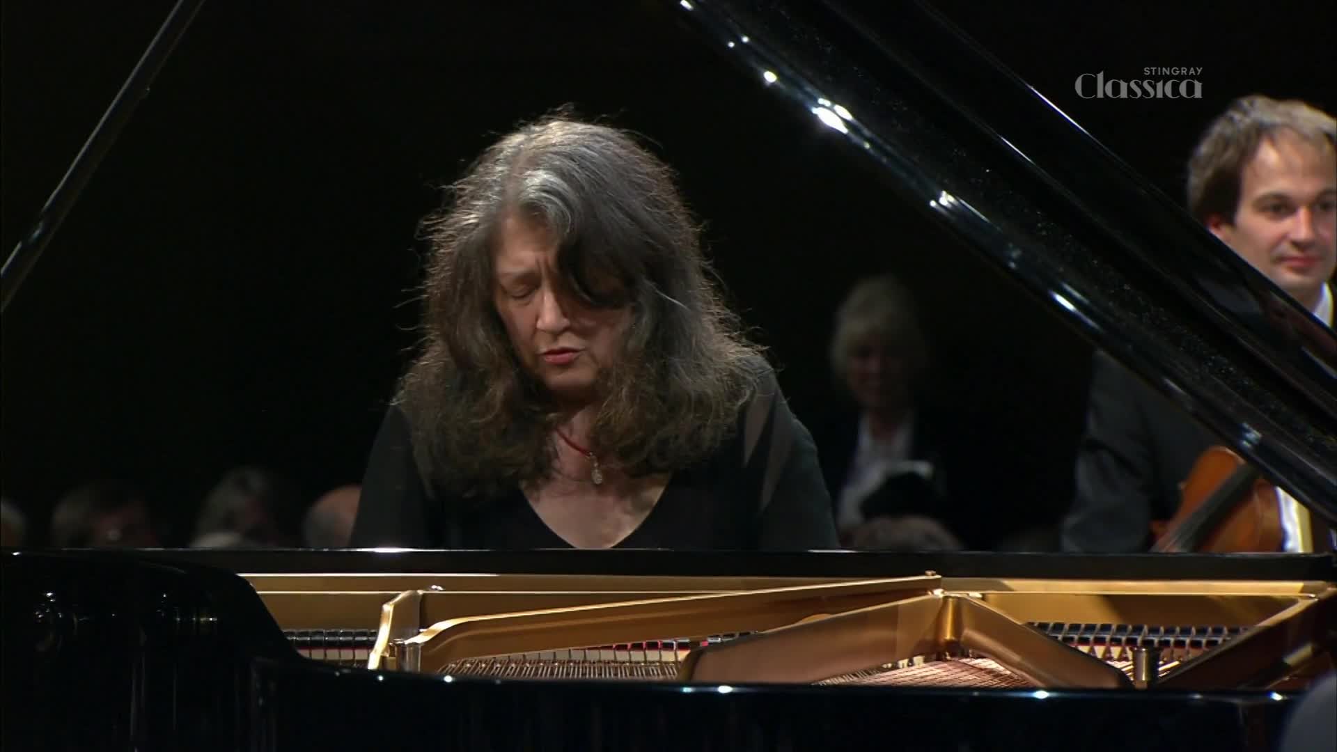 A woman with long, dark hair is playing the piano, her eyes closed in concentration. To her right, a man in a suit holds a cello, his gaze directed forward.