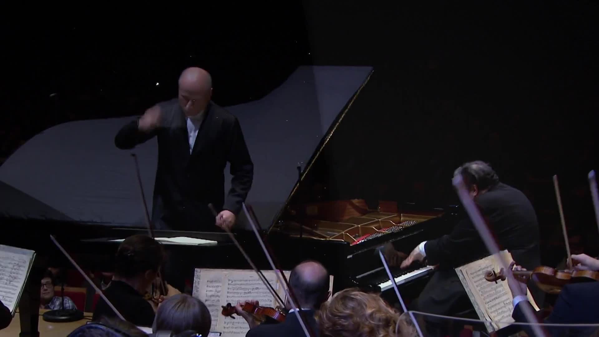 The conductor, a bald man in a black suit, gestures with his baton over a grand piano. A pianist, his back to us, plays the instrument while an orchestra surrounds them.