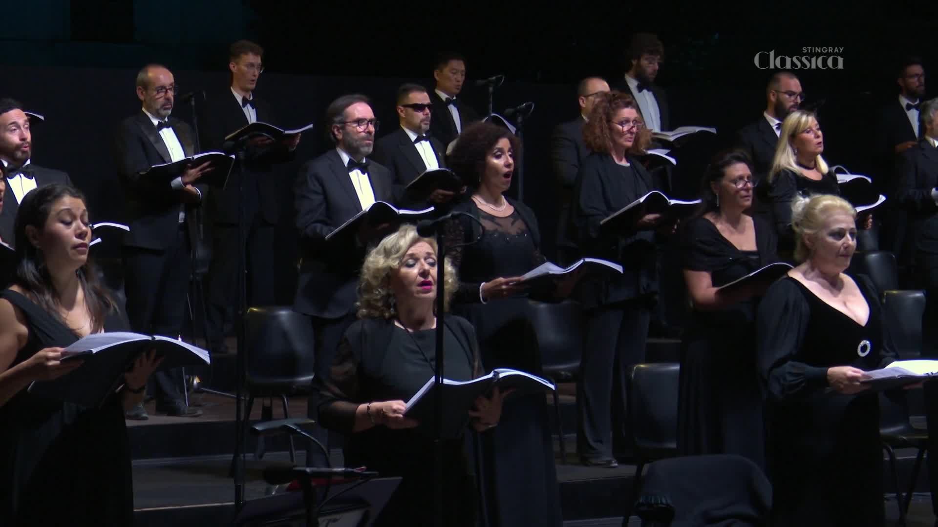 The choir, dressed in formal black, sings from their sheet music. A Stingray Classica logo appears on the screen, indicating this performance is being broadcast.