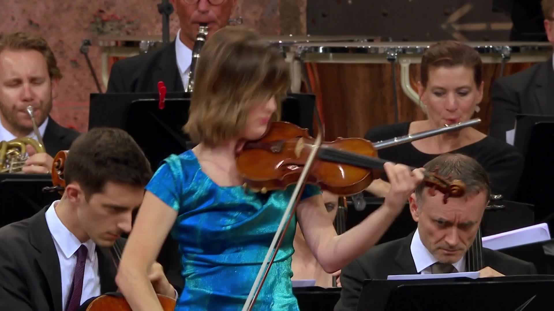 A young woman in a shimmering blue dress plays her violin, her bow moving across the strings. Behind her, an orchestra is in full swing, with a flutist and horn player visible.