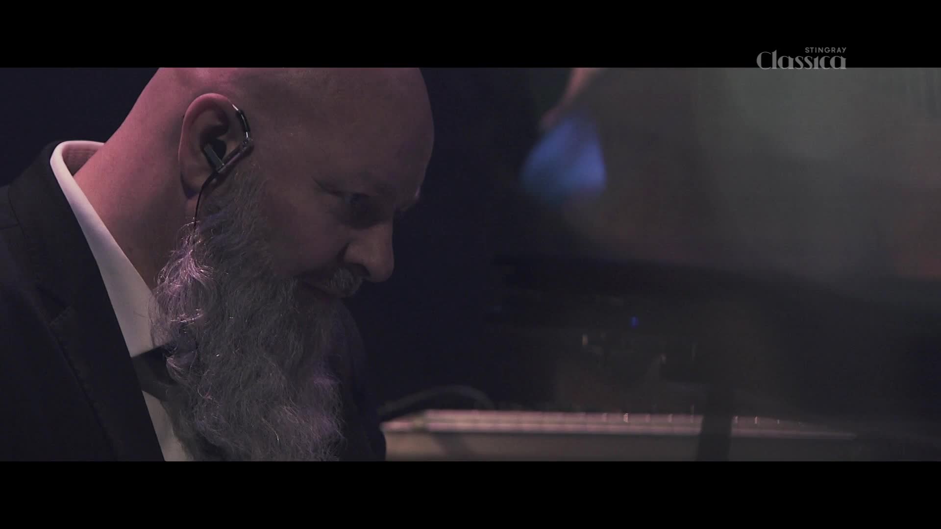 A man with a long grey beard and an earpiece focuses intently on the piano keys before him. The Stingray Classica logo is visible in the upper right corner.