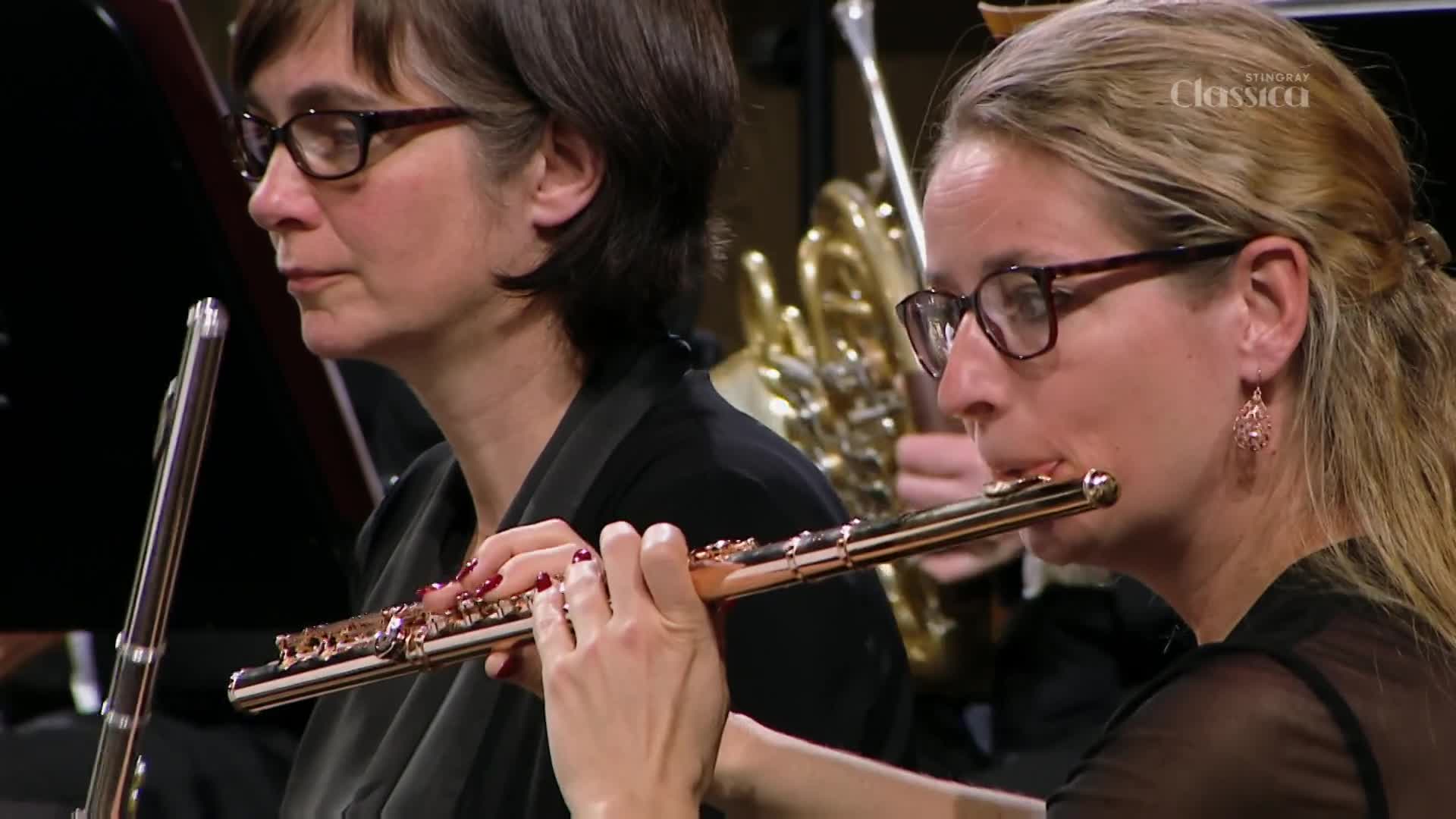 A woman with glasses plays a flute, her fingers moving across the keys. Beside her, another woman, also wearing glasses, holds her flute ready.