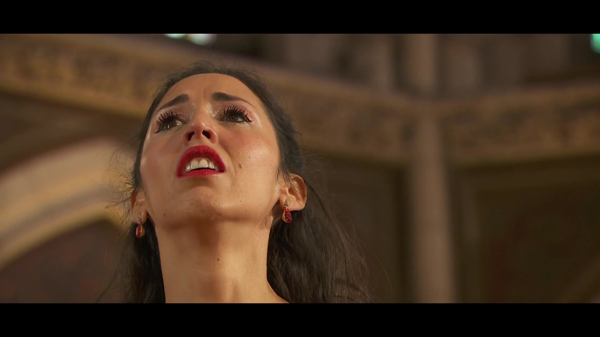 A woman with dark hair and bright red lipstick looks upward, her eyes wide and glistening. She wears small, ruby-colored earrings that dangle near her jawline.