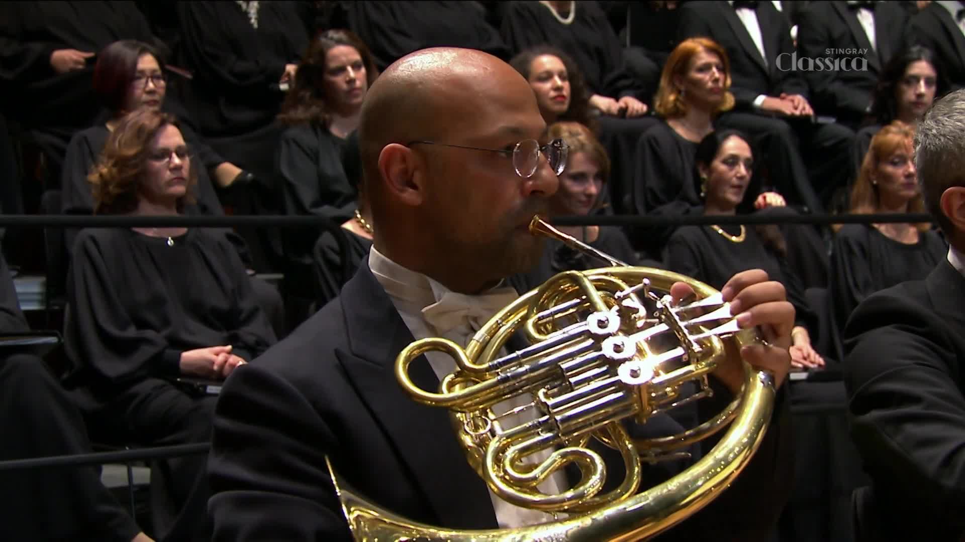 A man in a tuxedo plays a French horn, his cheeks puffed out as he blows into the mouthpiece. Behind him, a choir in black robes sits attentively, their faces turned towards the music.