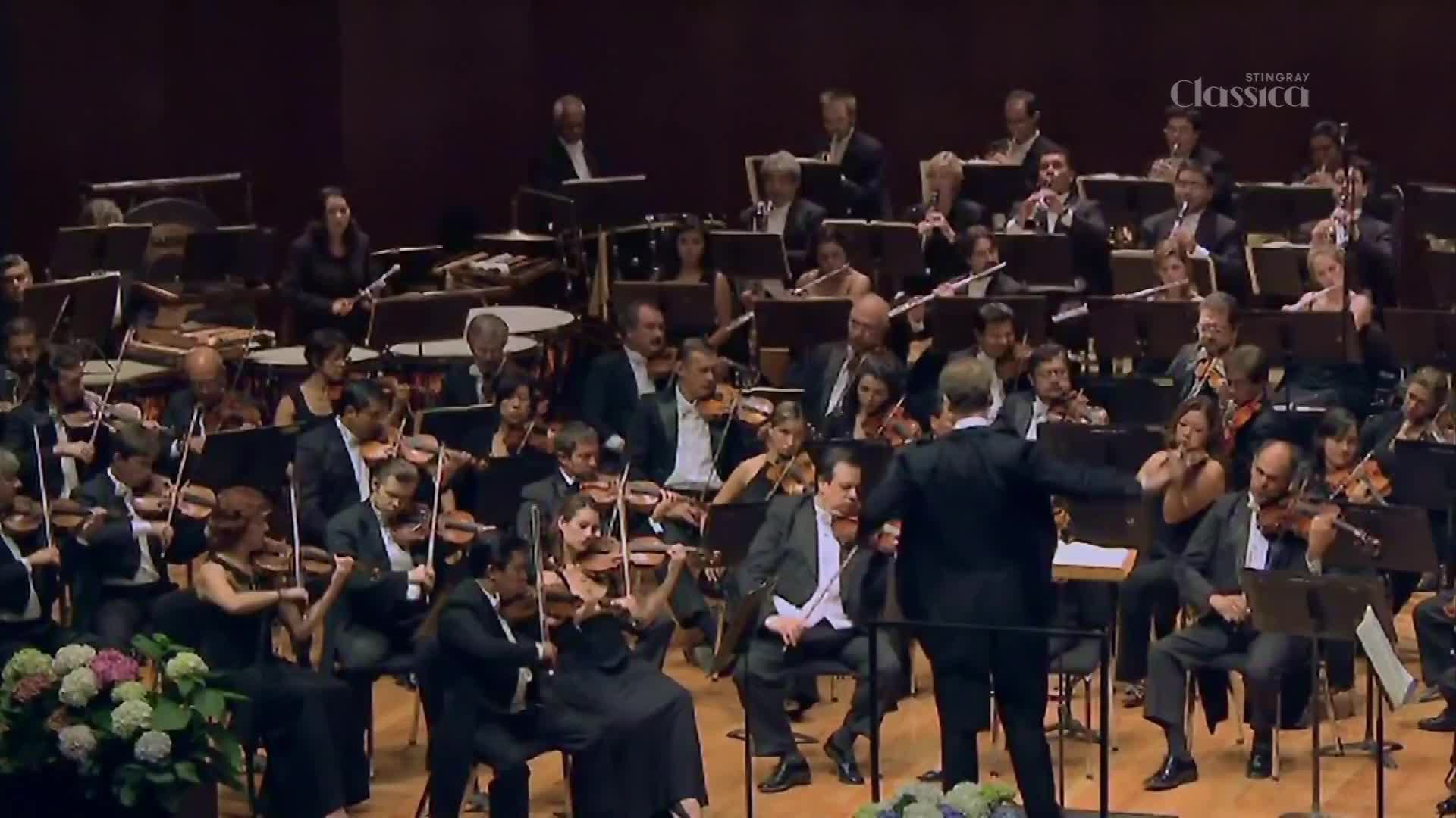 The conductor gestures with his left arm, leading a full orchestra in a performance. Violins are bowed across the stage, their players focused on the score.