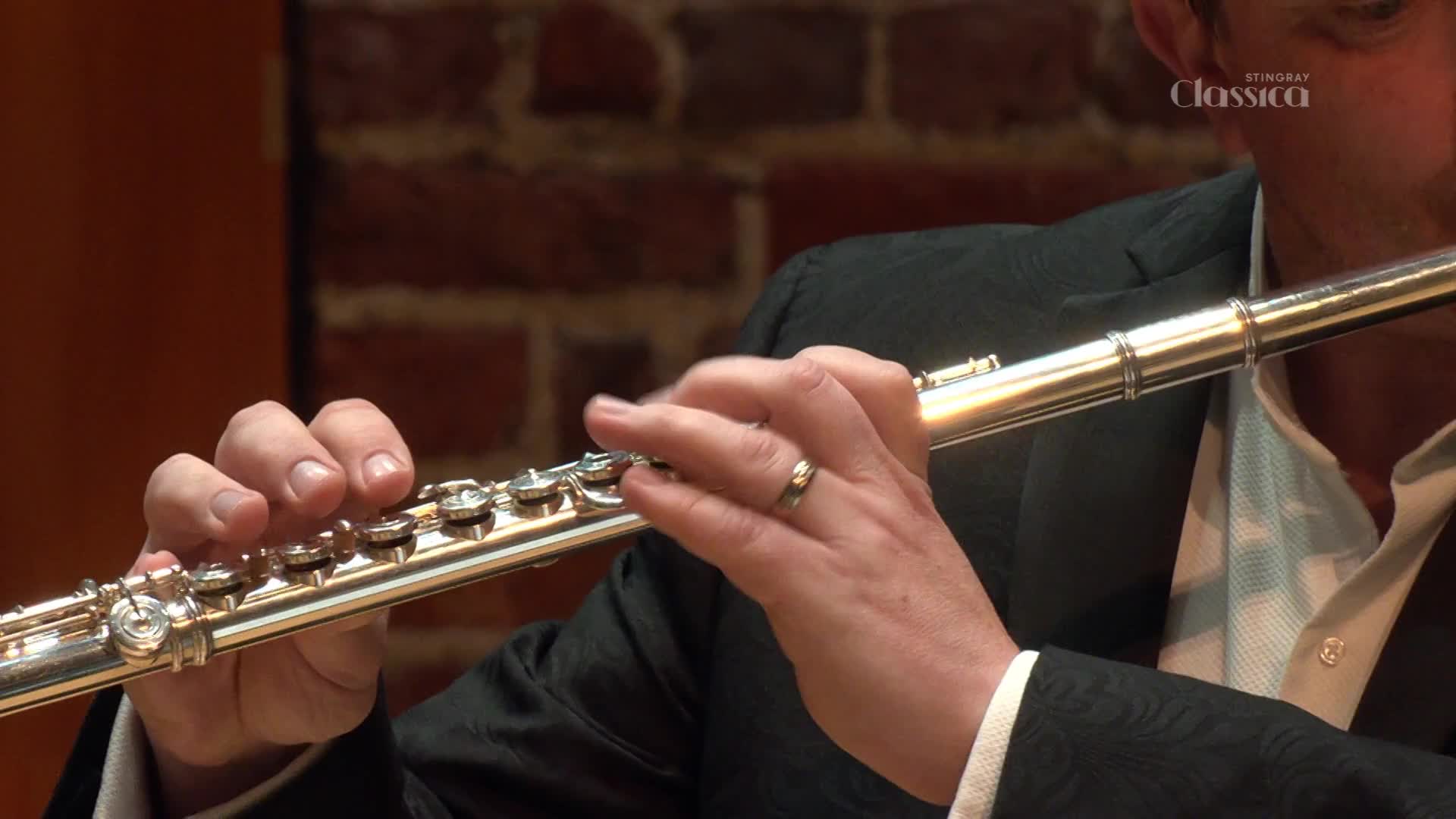 Fingers dance across the keys of a silver flute, the instrument held expertly. The musician, dressed in a dark suit, plays with focused intensity, his breath shaping the melody.