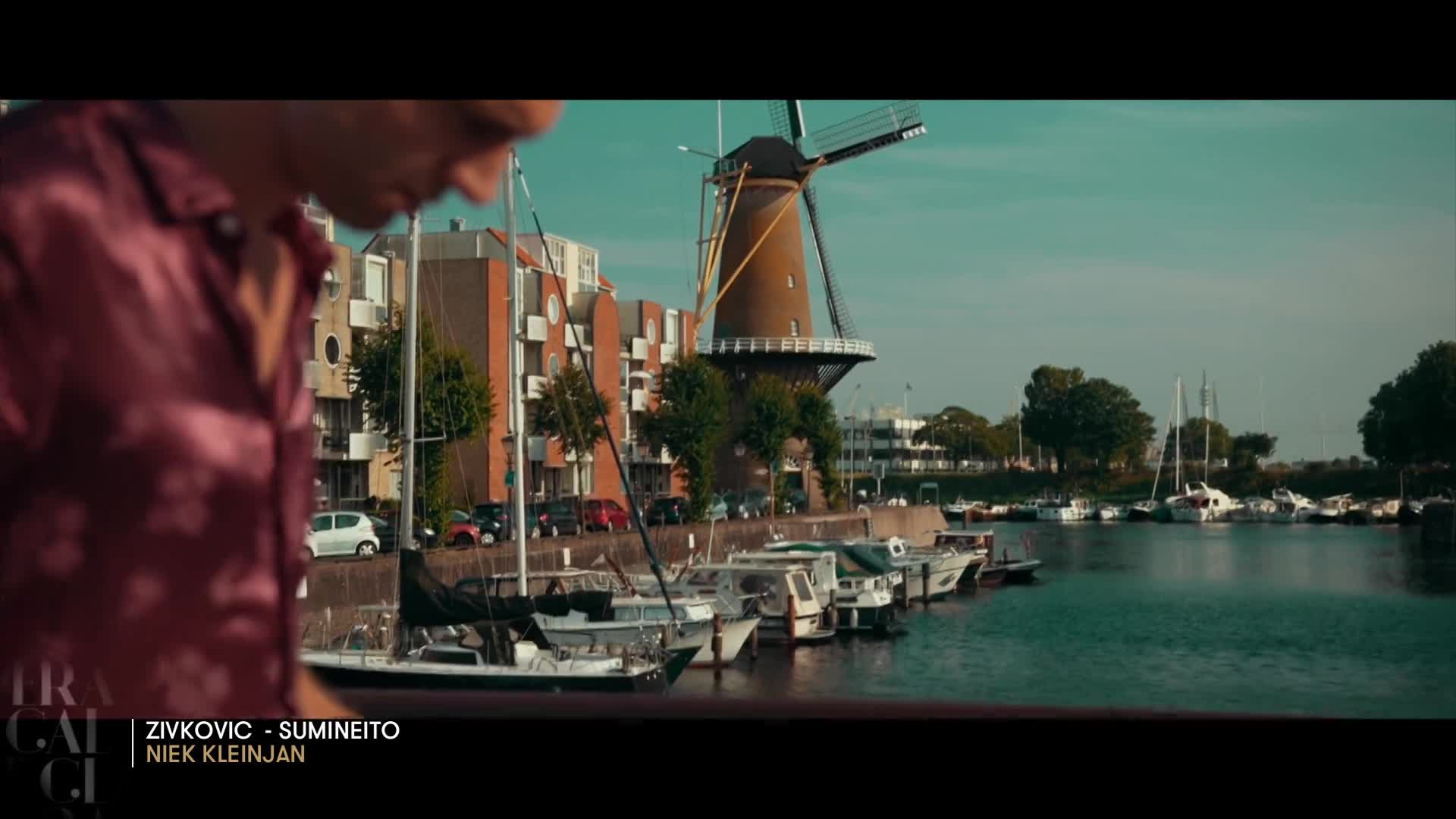 A classic Dutch windmill stands tall against a bright blue sky. Boats of various sizes are moored along the canal, their reflections shimmering on the water.