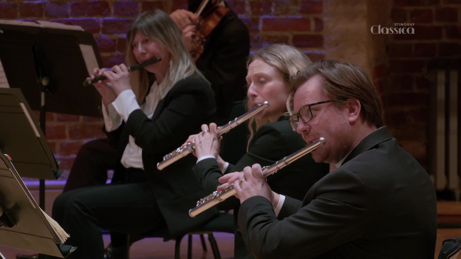 A trio of flutists plays in unison, their silver instruments catching the light. The Stingray Classica logo appears subtly in the background, hinting at a Canadian performance.